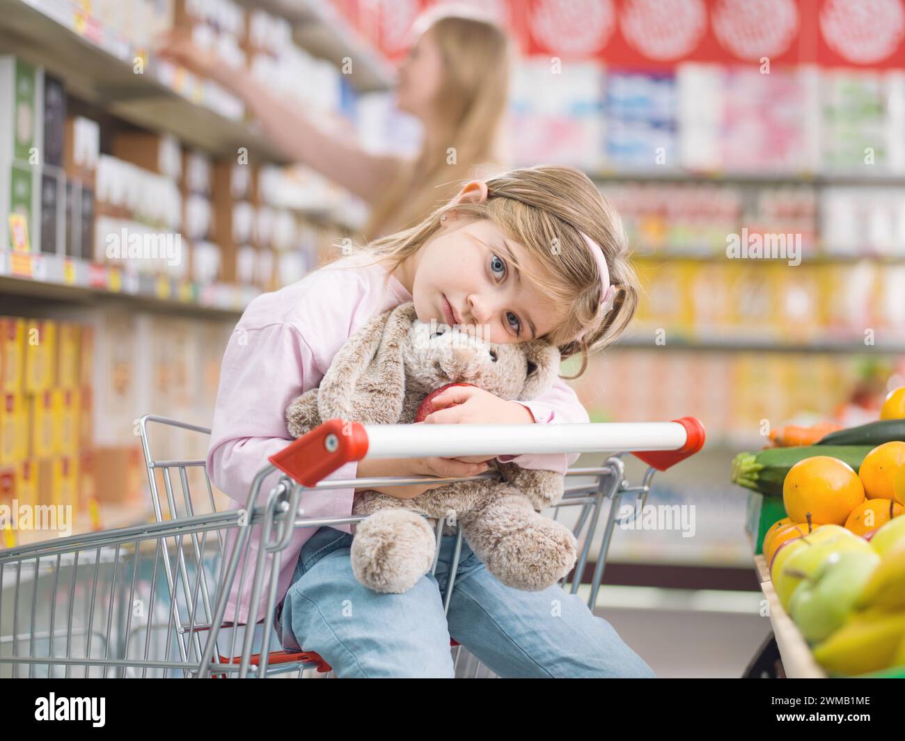 Niedliches gelangweiltes Kind, das auf einem Einkaufswagen sitzt und ihr Plüschtier umarmt, wartet auf ihre Mutter Stockfoto