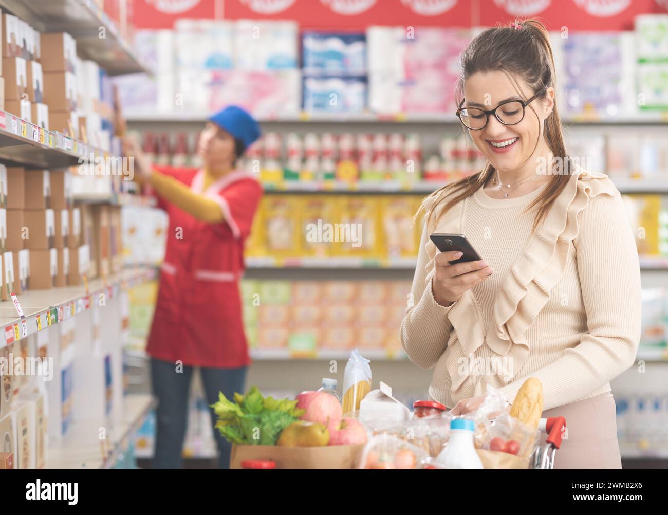 Lächelnde Frau, die Lebensmittel kauft und ein Smartphone im Supermarkt benutzt Stockfoto