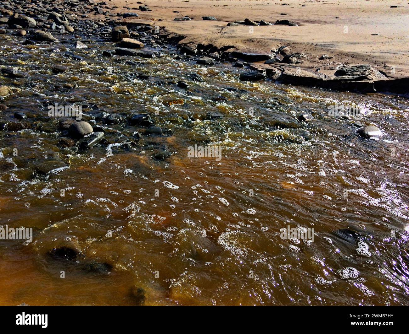 Ein Bach fließt über Felsen, sein braunes Wasser glitzert im Sonnenlicht, umgeben von einem felsigen Gelände. Stockfoto