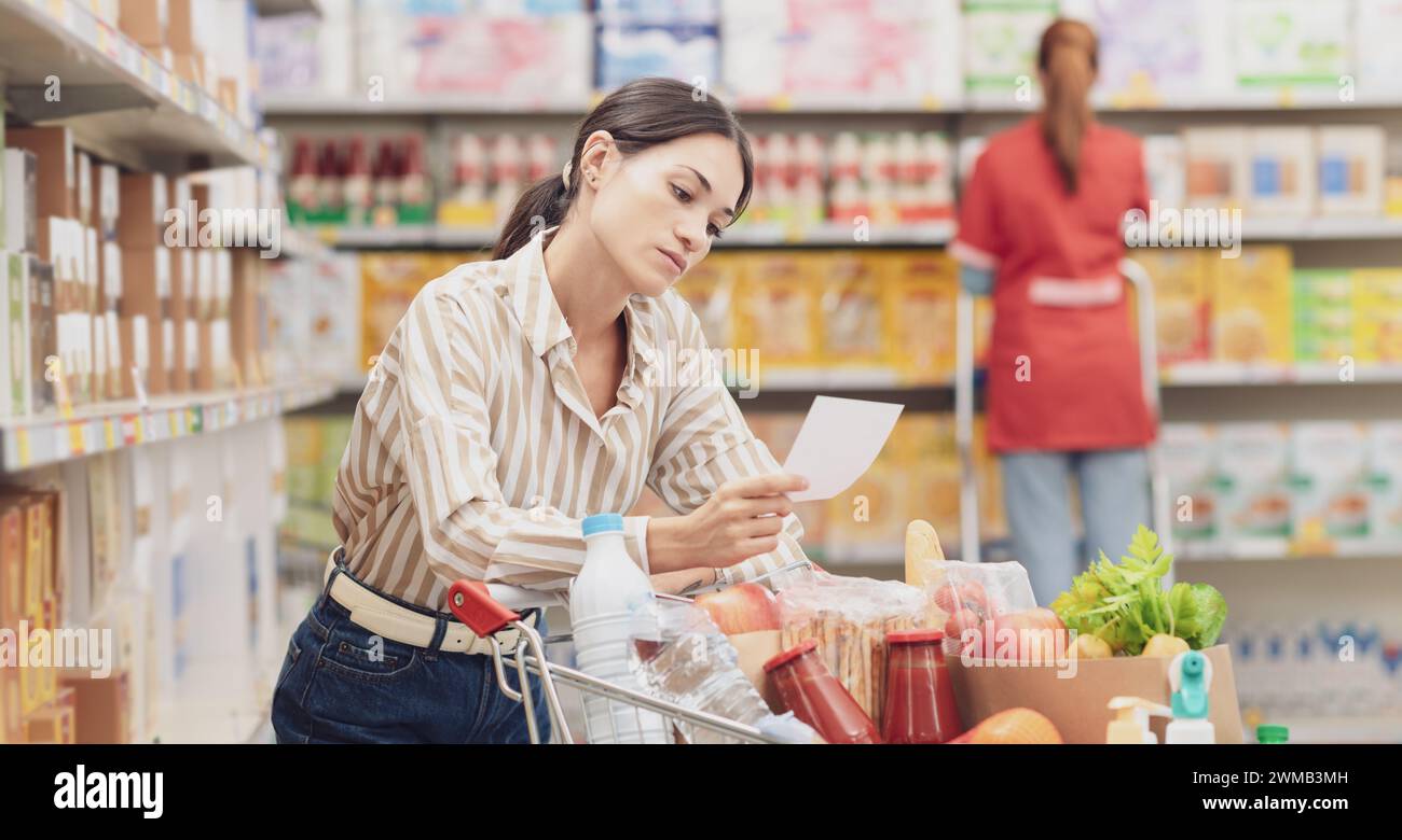 Junge nachdenkliche Frau mit vollem Einkaufswagen im Supermarkt, sie überprüft ihre Einkaufsliste, Lebensmitteleinkaufkonzept Stockfoto