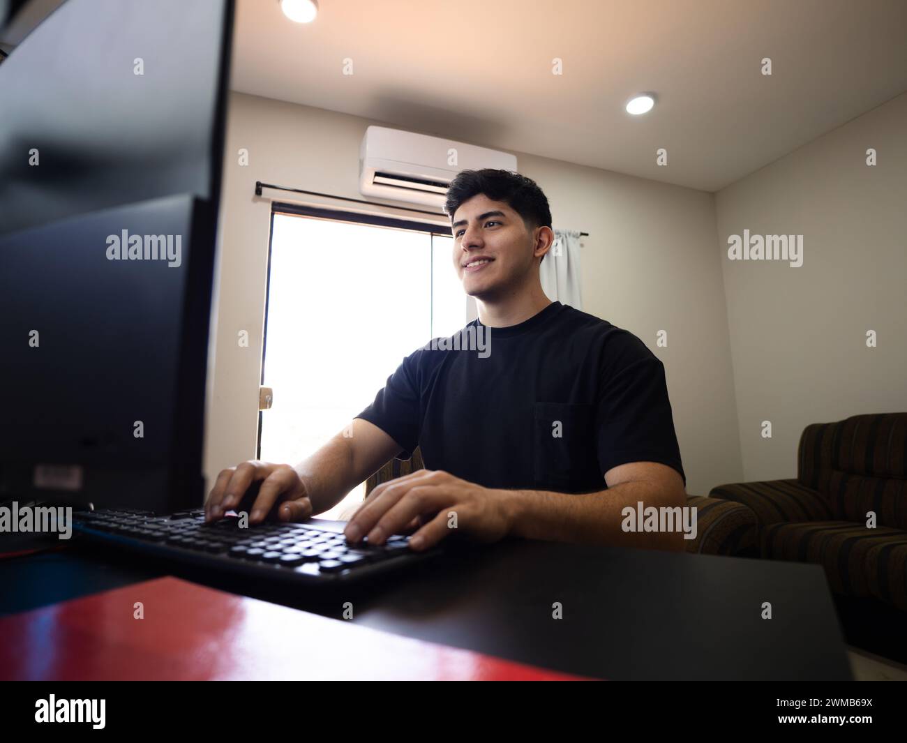 Ein lächelnder, engagierter junger Mann vor dem Computer, der Aufgaben schreibt, Projekte plant und in die Welt der Fernarbeit eintaucht Stockfoto
