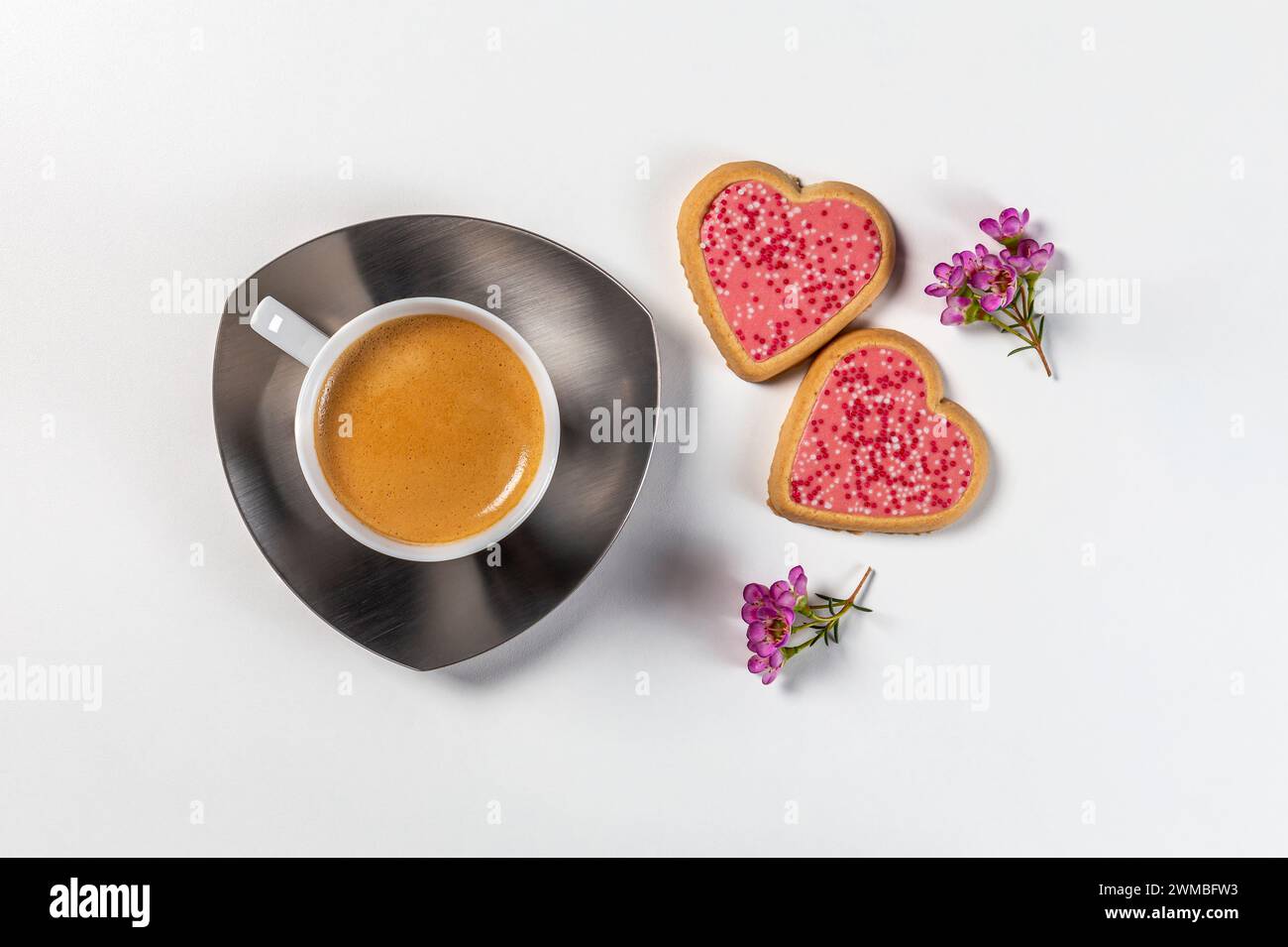 Eine Tasse Kaffee auf einer Untertasse mit zwei herzförmigen Keksen in rosafarbener Glasur und Blütenzweigen. Draufsicht auf weißem Hintergrund. Stockfoto