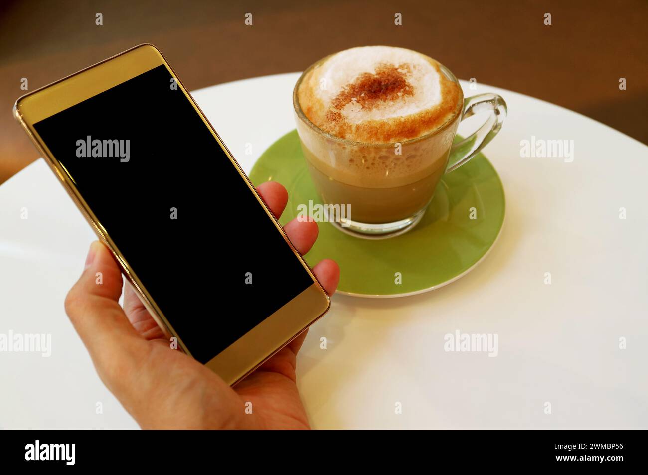 Hand hält Handy mit leerem Bildschirm und einer Tasse Cappuccino im Hintergrund Stockfoto