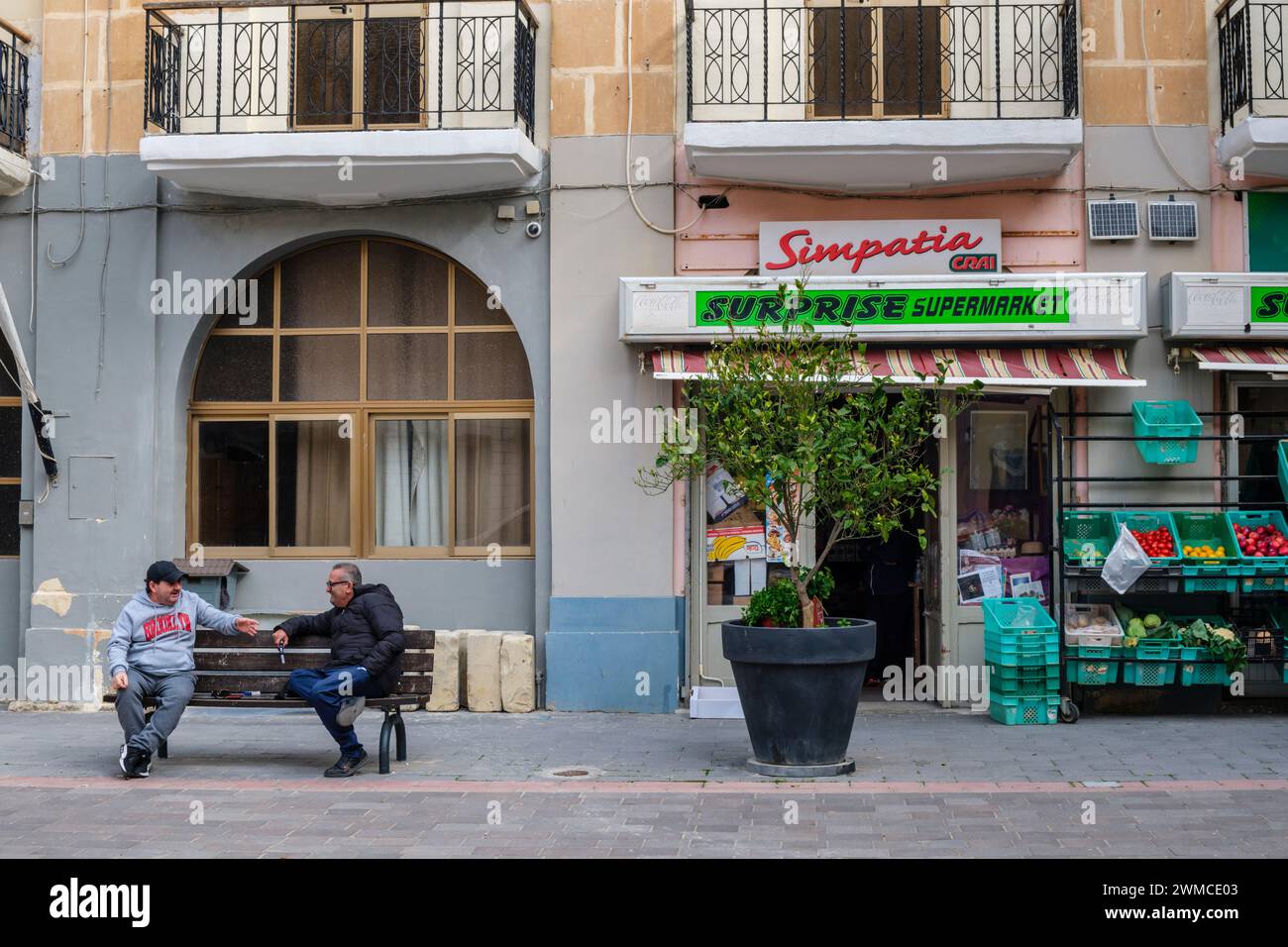Zwei Männer unterhalten sich auf einer Bank vor einem Supermarkt in Senglea, Valletta, Malta Stockfoto