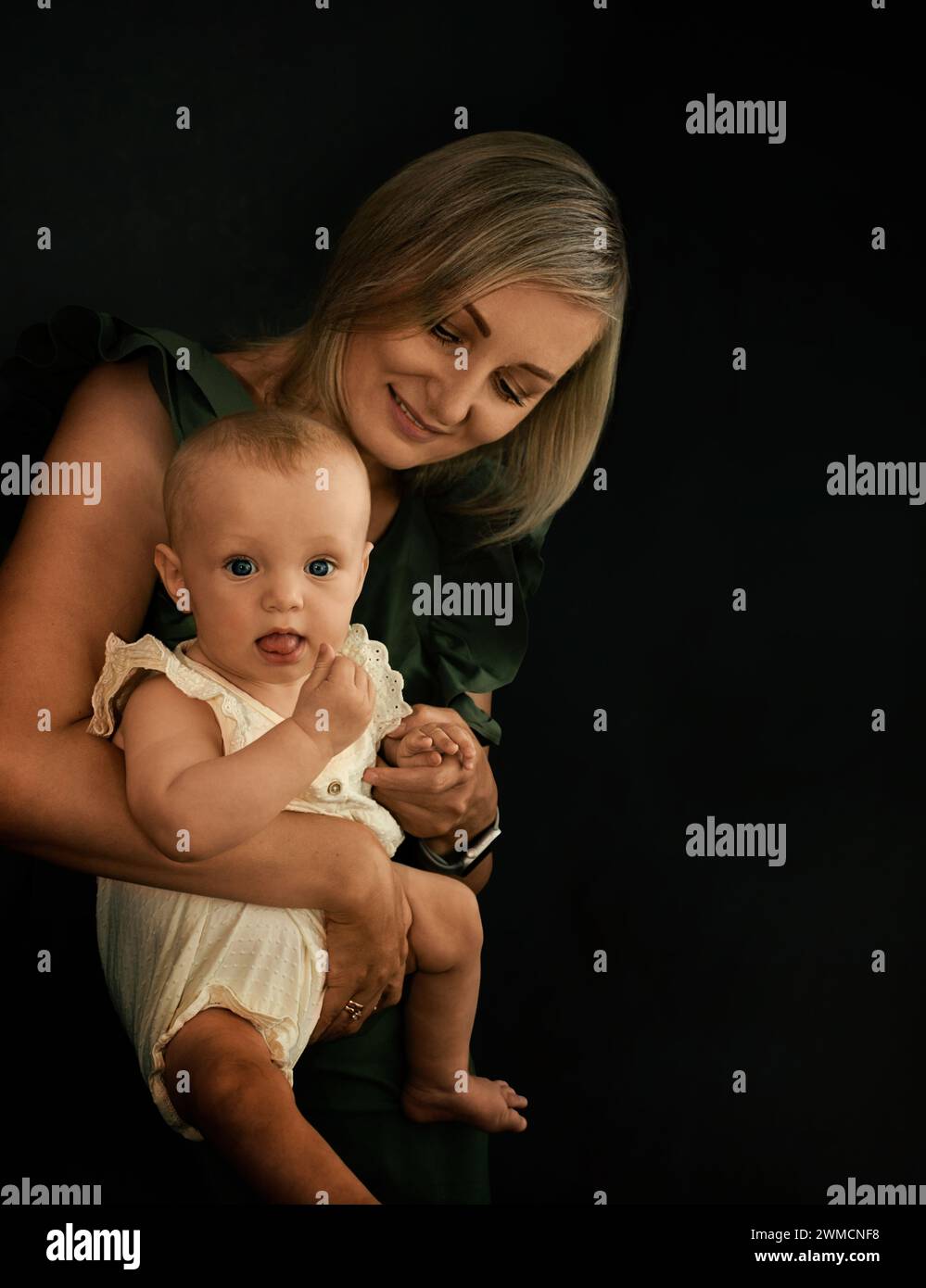 Junge blonde Mutter, die ein Baby in den Armen auf schwarzem Hintergrund in der Nähe des Fensters hält Stockfoto