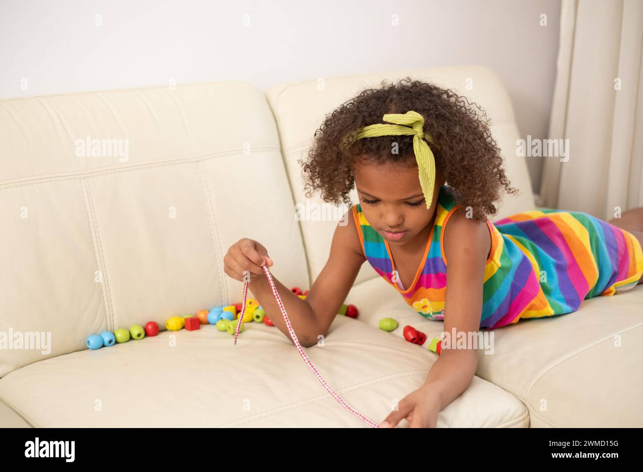 Ein Mulatto-Kind auf der Couch, das sich auf Perlen konzentriert, eine Kette, ein gelbes Haarreif und ein gestreiftes Regenbogenkleid. Ein Moment der Kreativität und des Lernens zu Hause, Bildung Stockfoto