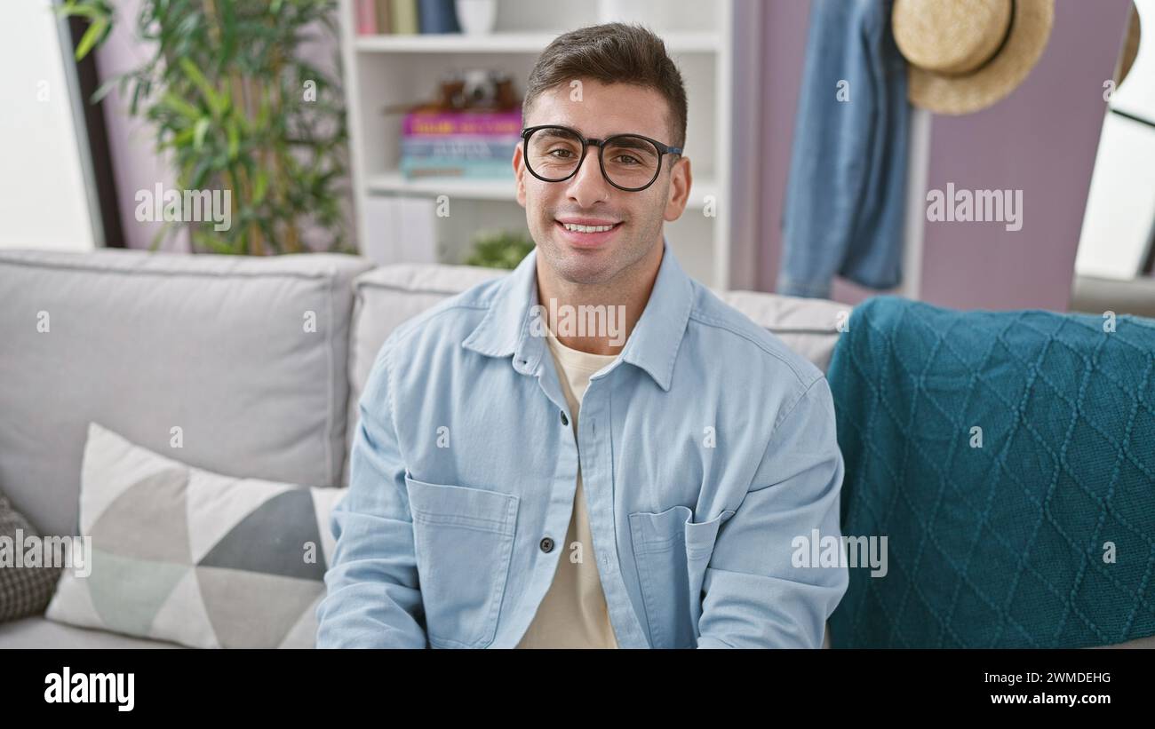 Fröhlicher, gutaussehender junger hispanischer Mann mit einem selbstbewussten Lächeln, der sich bequem auf dem Sofa im Wohnzimmer zu Hause entspannt und dabei die Gemütlichkeit im Innenraum genießt Stockfoto