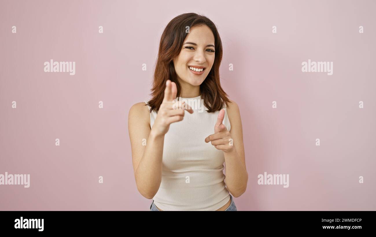 Eine junge hispanische Frau mit modernem Haar lächelt und zeigt vor einem rosa isolierten Hintergrund auf die Kamera, die Positivität und Engagements zeigt Stockfoto