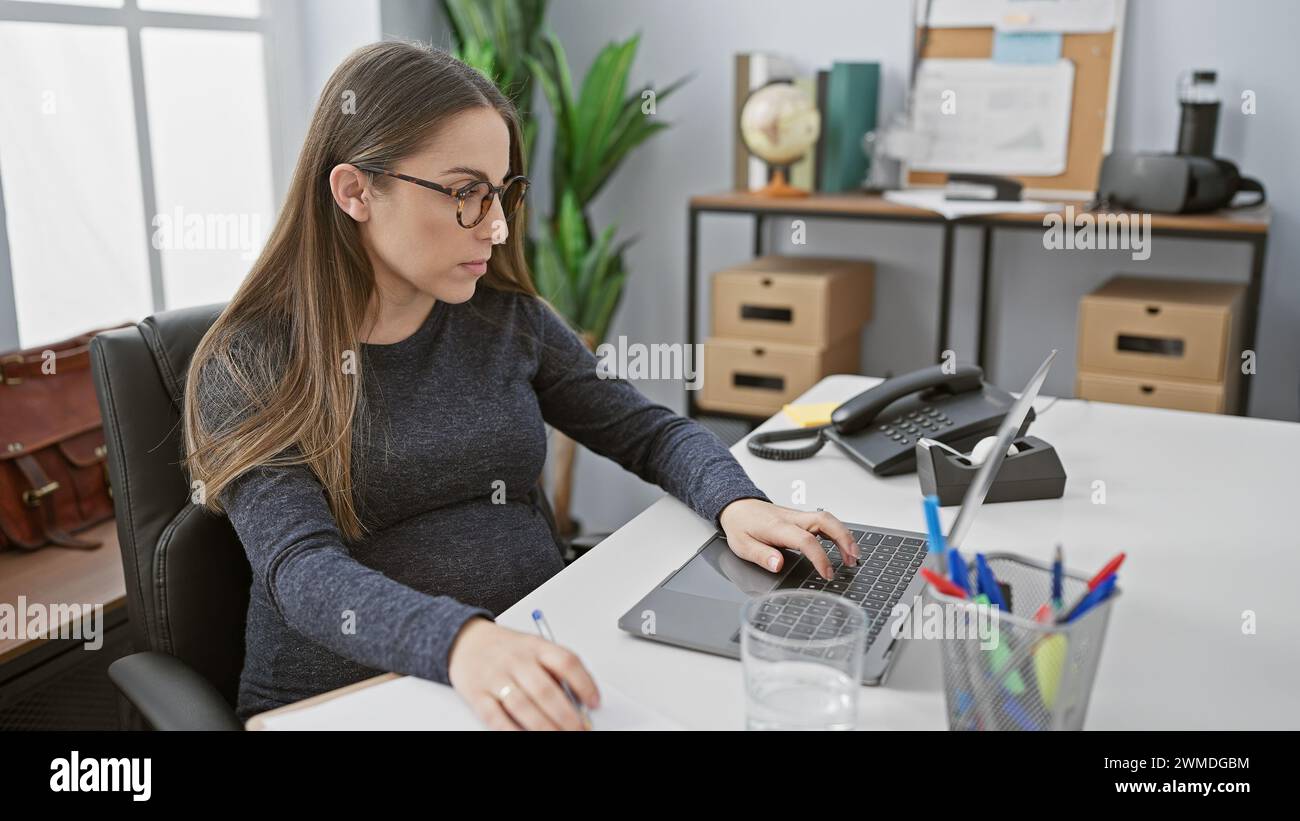 Eine schwangere hispanische Frau arbeitet an ihrem Schreibtisch und erinnert an Themen wie Professionalität, Mutterschaft und Arbeitsumfeld in Innenräumen. Stockfoto
