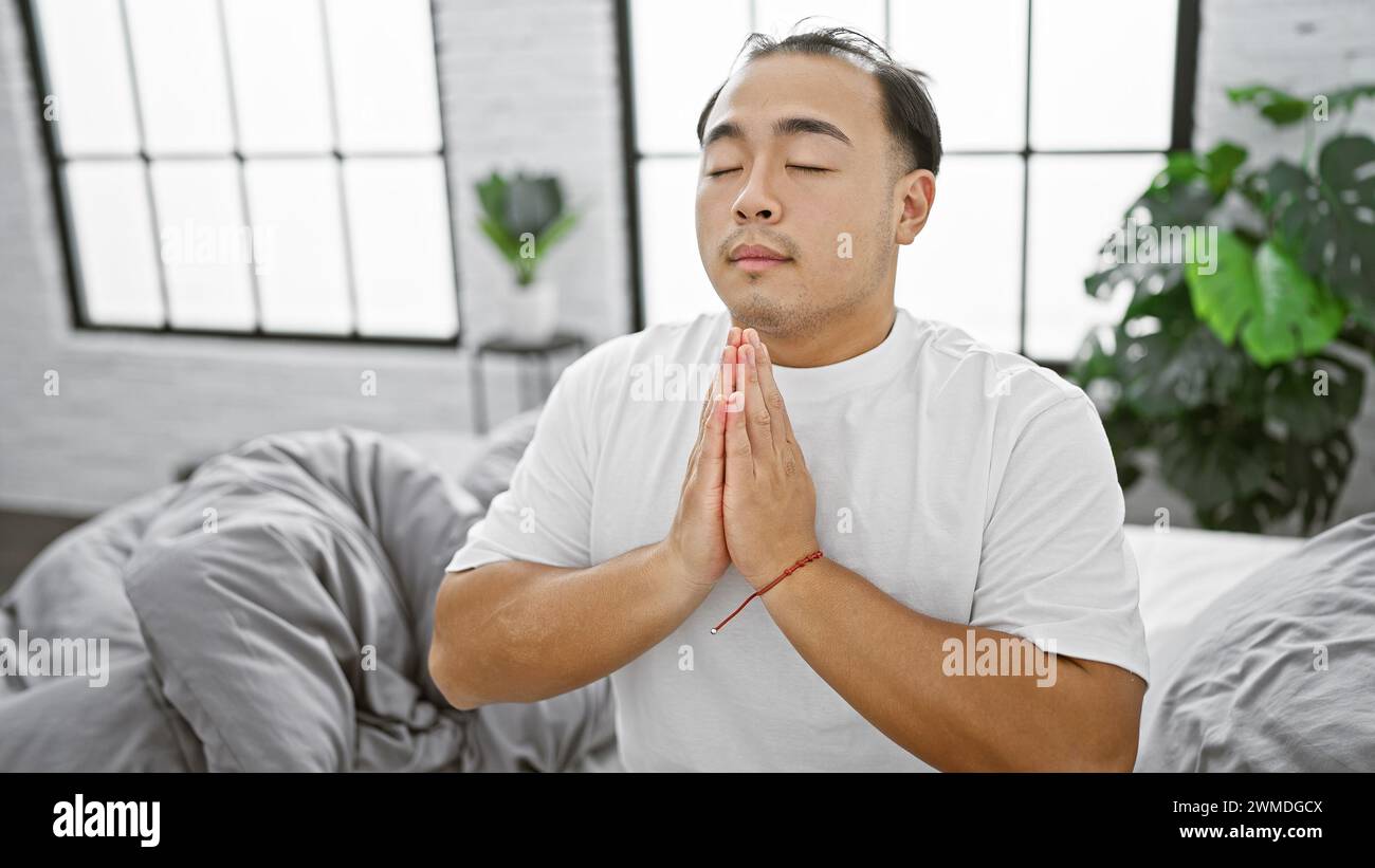 Ein junger, gutaussehender chinesischer Mann, der seinen Glauben an jesus im Gebet zum Ausdruck bringt, während er sich auf einem bequemen Bett in der gemütlichen Inneneinrichtung entspannt Stockfoto