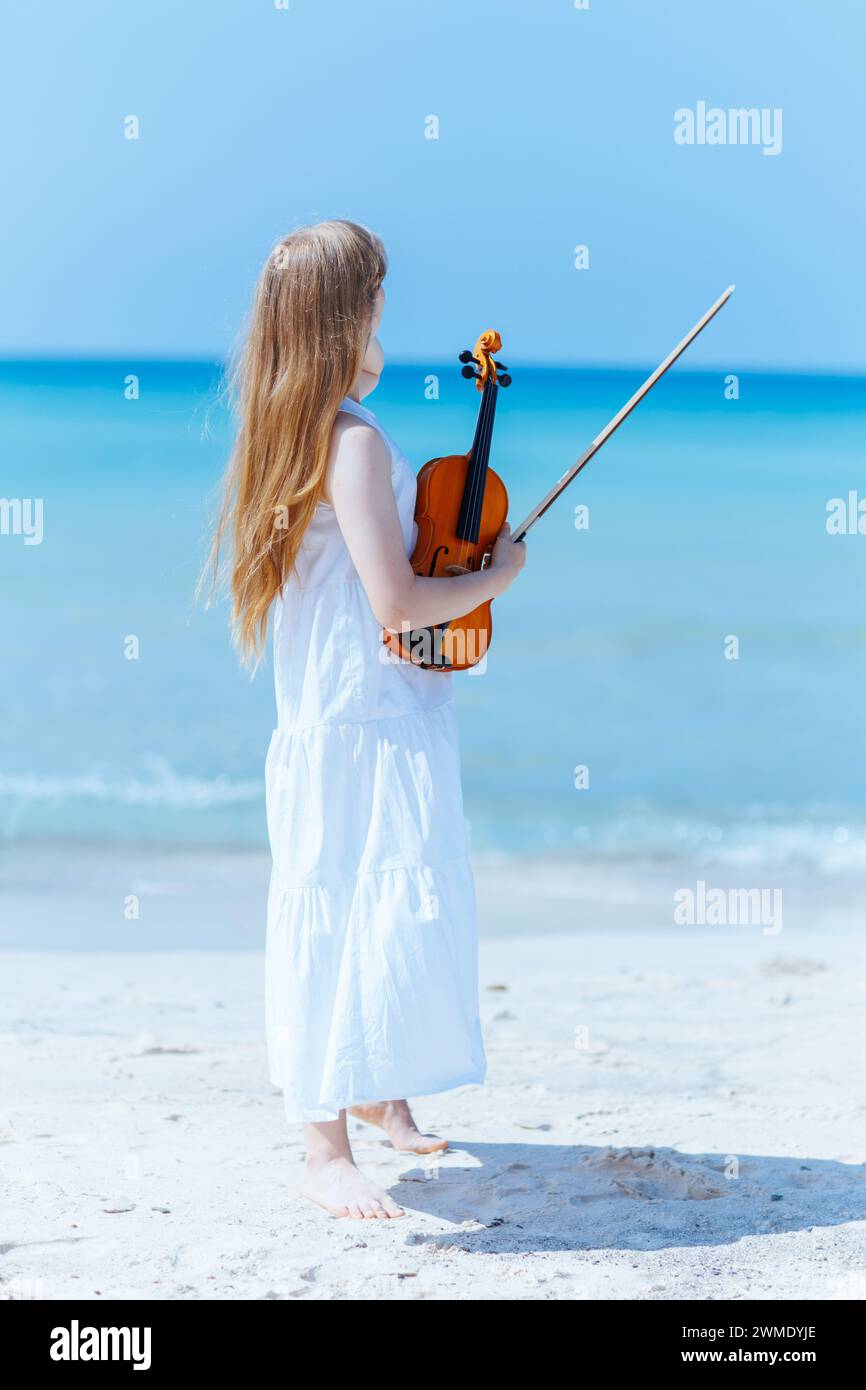 Porträt eines entspannten modernen Teenagers in weißem Kleid an der Meeresküste mit Geige. Stockfoto