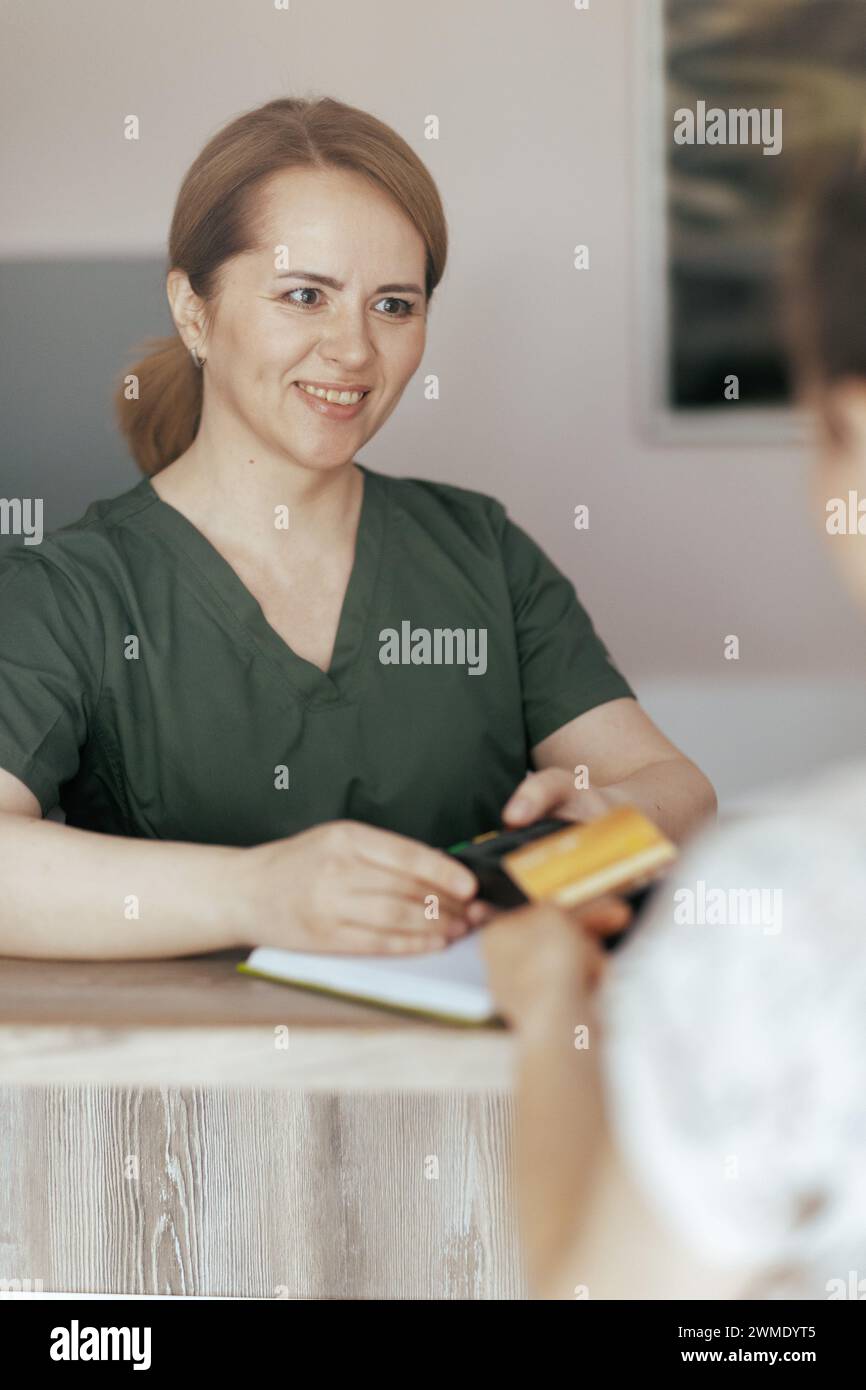 Gesundheitszeit. Lächelnde Massage-Therapeutin im Massageschrank, der Kunde akzeptiert Kreditkartenzahlungen über das Terminal an der Rezeption. Stockfoto