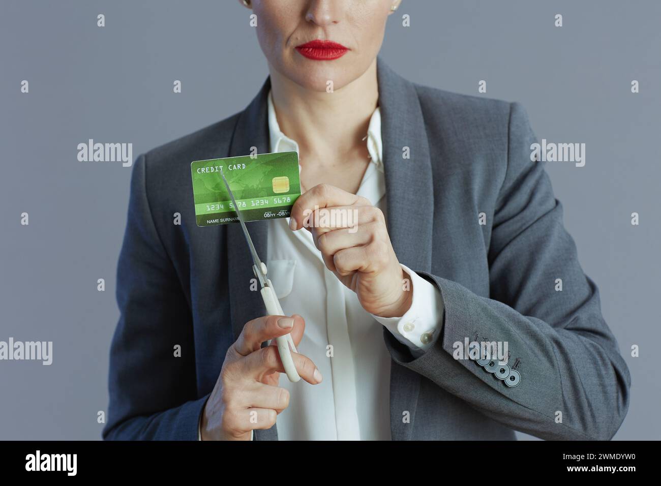 Nahaufnahme einer Mitarbeiterin in grauem Anzug mit Kreditkarte und Schere vor grauem Hintergrund. Stockfoto
