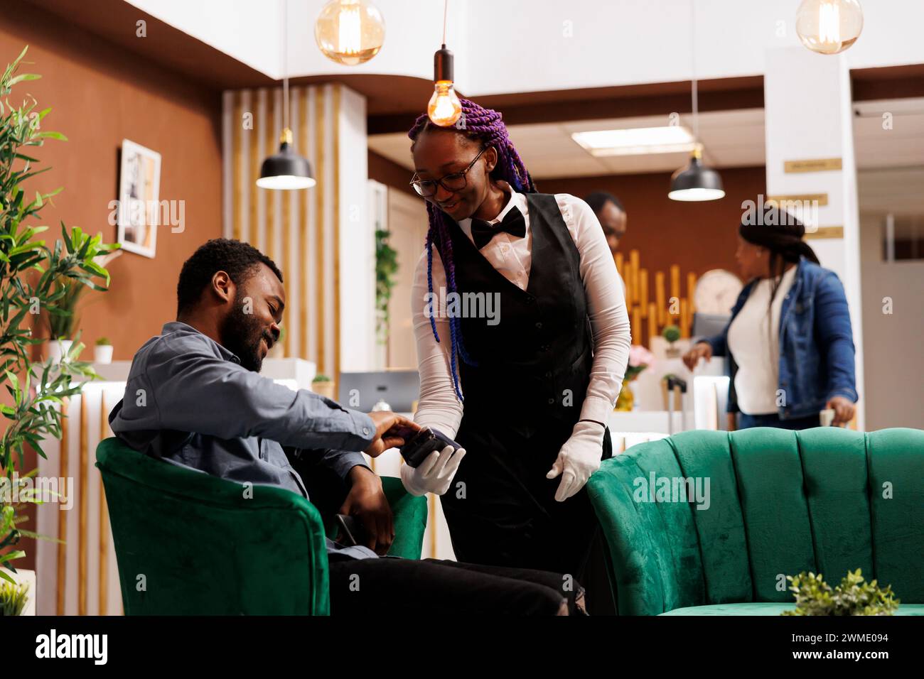 Lächelnder afroamerikanischer Tourist, der eine Kreditkarte bezahlt, während er sich in der Lounge des Hotels erholt. Freundliche weibliche Kellnerin mit Pos Terminal, die Gäste in der Hotellobby bedient Stockfoto