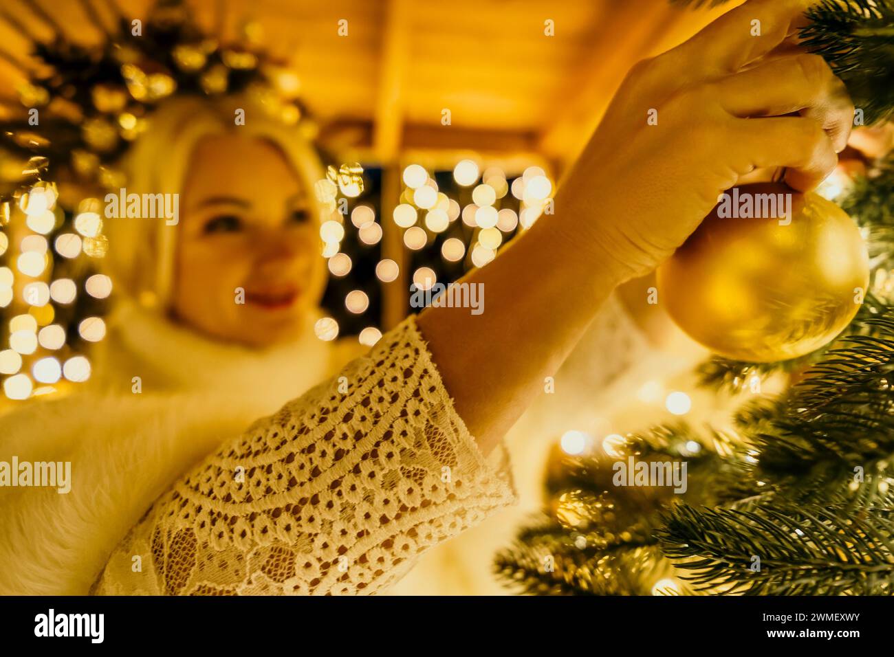 Eine blonde Frau in weißem Kleid und eine Krone aus goldenen Ornamenten schmücken den Weihnachtsbaum mit goldenen Ornamenten und Lichtern. Der Baum ist mit Gold verziert Stockfoto