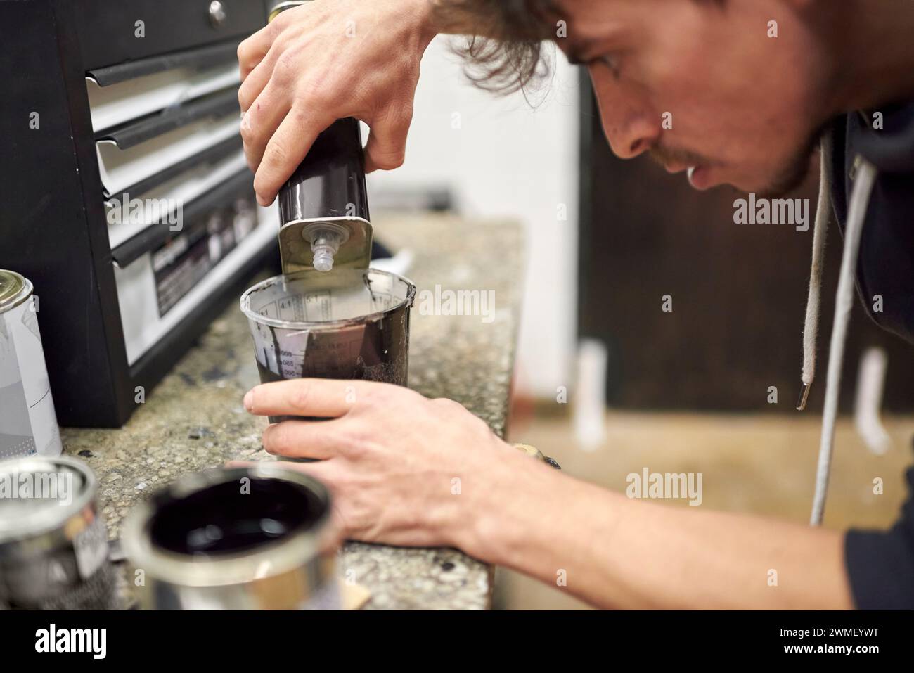 Junger hispanischer Mann, der dünner in eine Tasse schwarze Farbe auf einer Werkstattarbeitsplatte gießt. Echte Menschen bei der Arbeit. Stockfoto