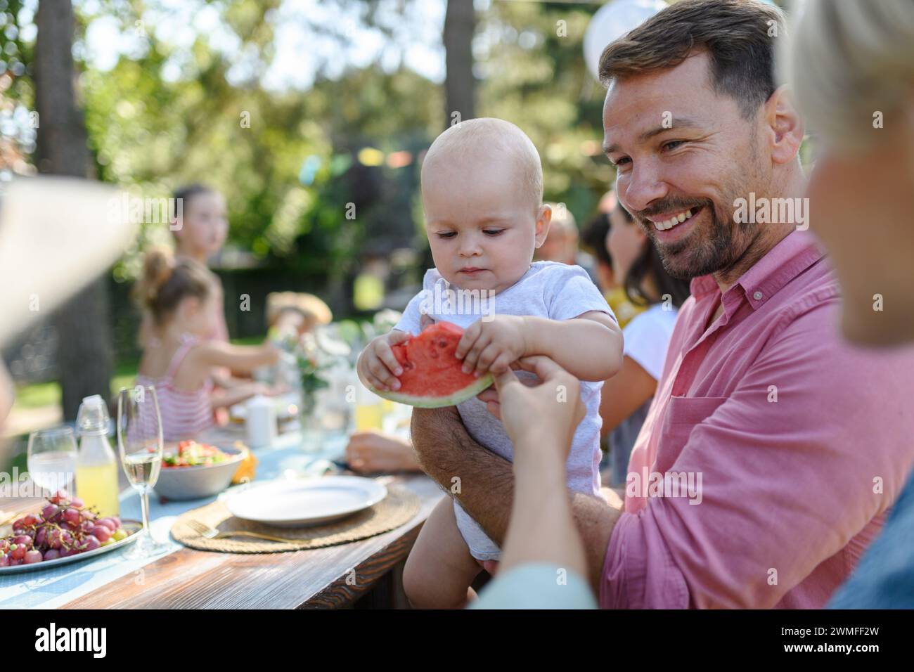 Süßes Baby isst Wassermelone auf einer Gartenparty im Sommer. Dad hält den Jungen in den Armen und lächelt. Stockfoto