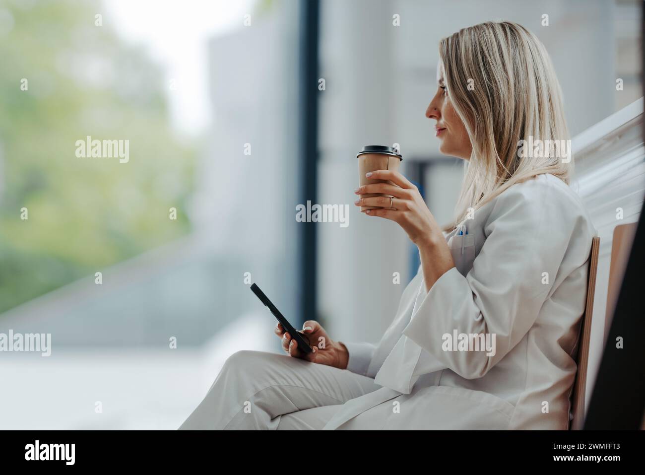 Die schöne Ärztin macht während ihrer Arbeitsschicht im Krankenhaus eine Pause, trinkt Kaffee und scrollt auf dem Smartphone. Seitenansicht. Stockfoto