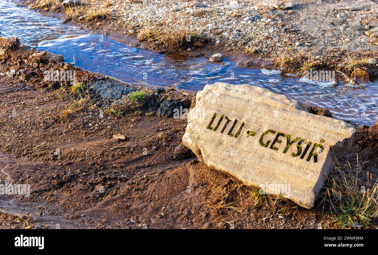 Blick von oben auf das Betonsteinstück mit der Inschrift Litli Geysir am Ufer eines künstlichen kleinen Baches mit Quellwasser und Blasen am Boden bei Tageslicht. Die Stockfoto