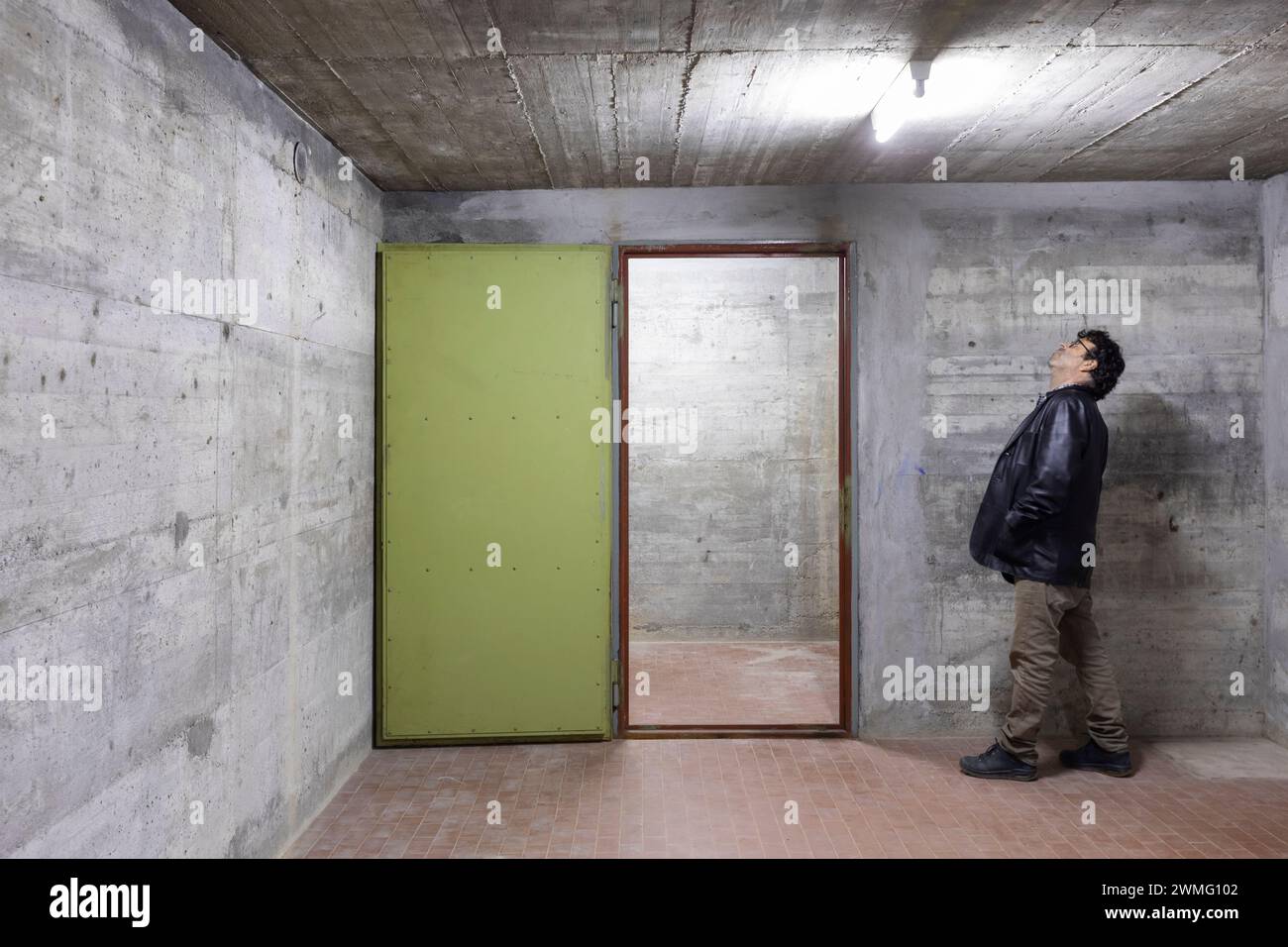 Ein Mann, der nach oben schaut, versucht zu verstehen, was außerhalb des Bunkers passiert, wenn die Sicherheitstür des Tresors offen ist. Szene beleuchtet von einem weißen Neon la Stockfoto