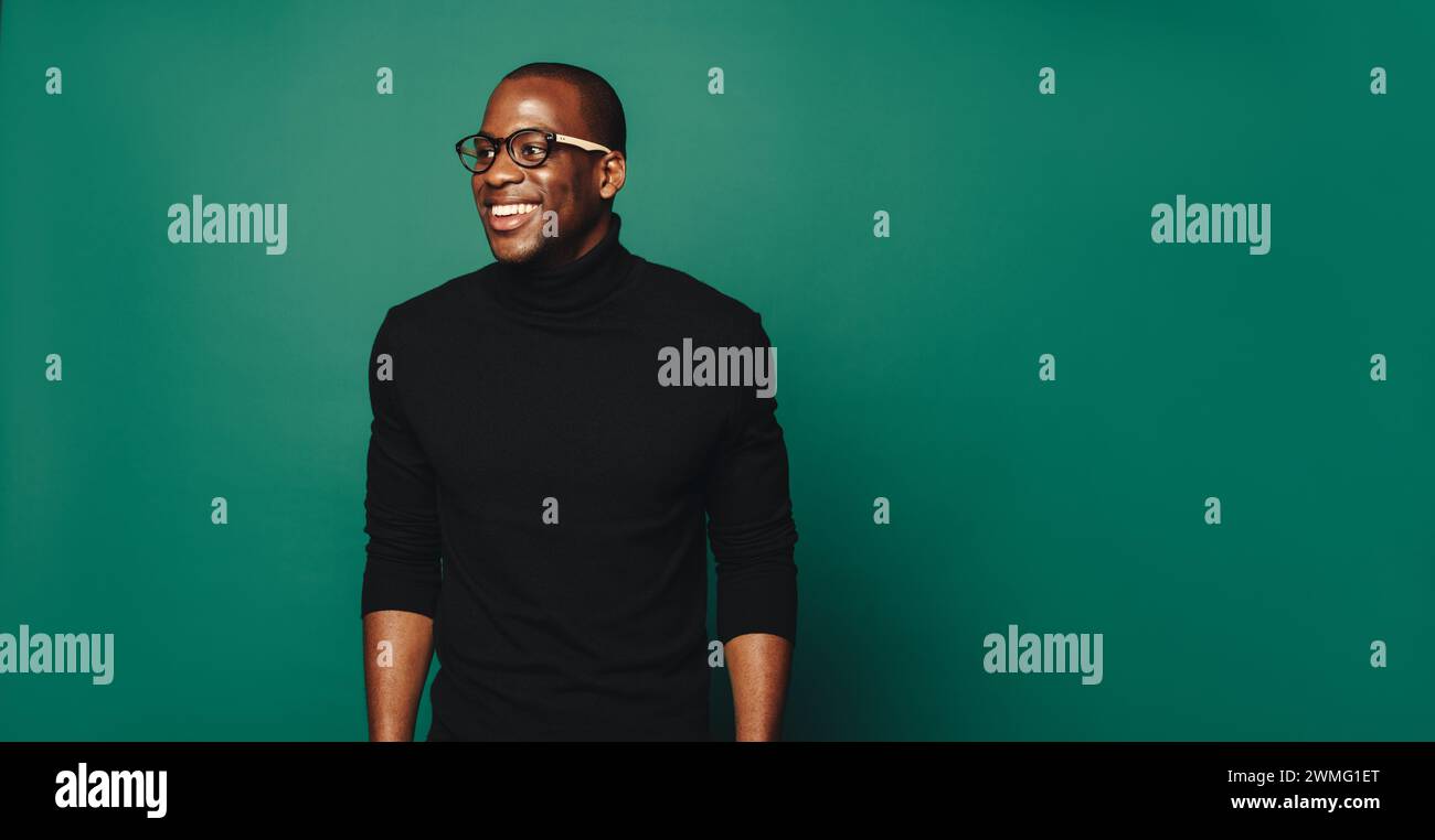 Ein selbstbewusster und stilvoller junger Mann steht in einem Studio vor einem trendigen grünen Hintergrund. Er trägt ein fröhliches Lächeln und einen lässigen, aber eleganten Pullover Stockfoto