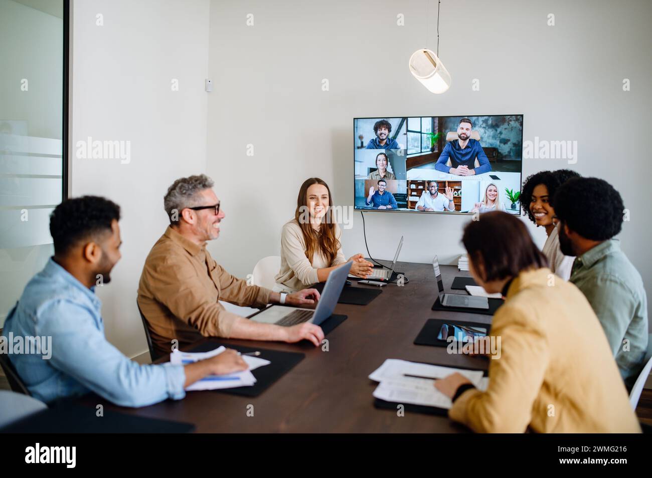 Ein lebhaftes und produktives Team-Meeting wird aufgezeichnet, bei dem Kollegen über einen großen Bildschirm miteinander verbunden sind und eine Mischung aus persönlicher und Remote-Zusammenarbeit darstellen. Konzept für virtuelle Besprechungen Stockfoto