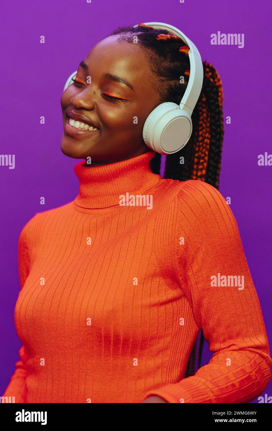 Glückliche Frau mit bunten zweifarbigen Zöpfen. Sie steht vor einem leuchtend violetten Hintergrund und trägt legere Kleidung und Kopfhörer. Ihr modernes Make-up A Stockfoto
