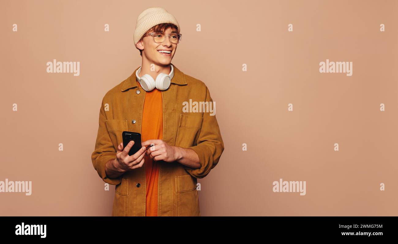 Happy man trägt lässige Denim-Kleidung und steht vor pfirsichfarbenem Hintergrund in einem Studio. Er hält ein Smartphone, nutzt eine mobile App und sendet SMS. Vibran Stockfoto