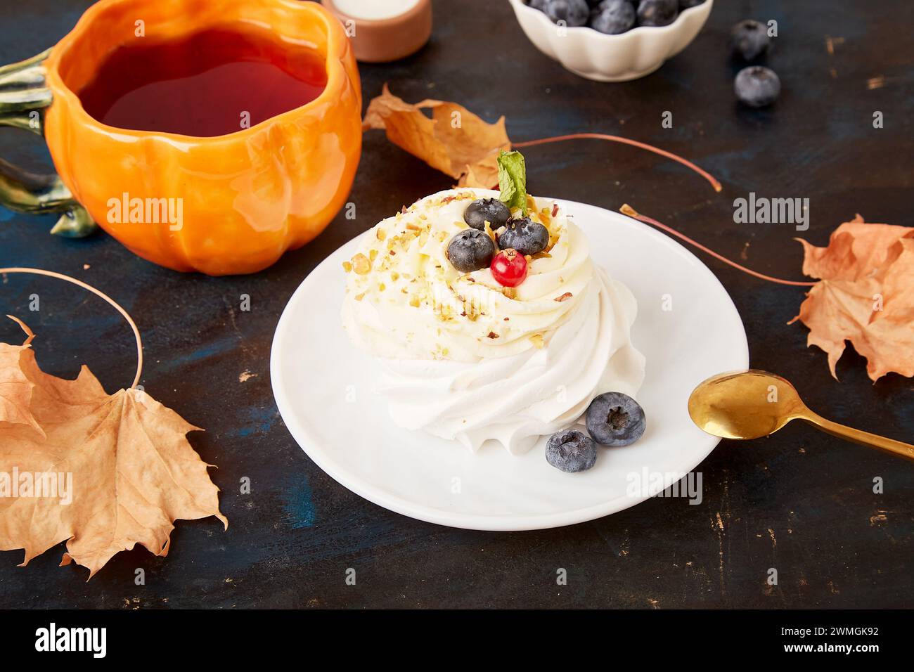 Genießen Sie herbstliche Vibes mit Pavlova und einem Kürbis-Teetasse unter trockenen Blättern auf rustikalem Hintergrund. Stockfoto