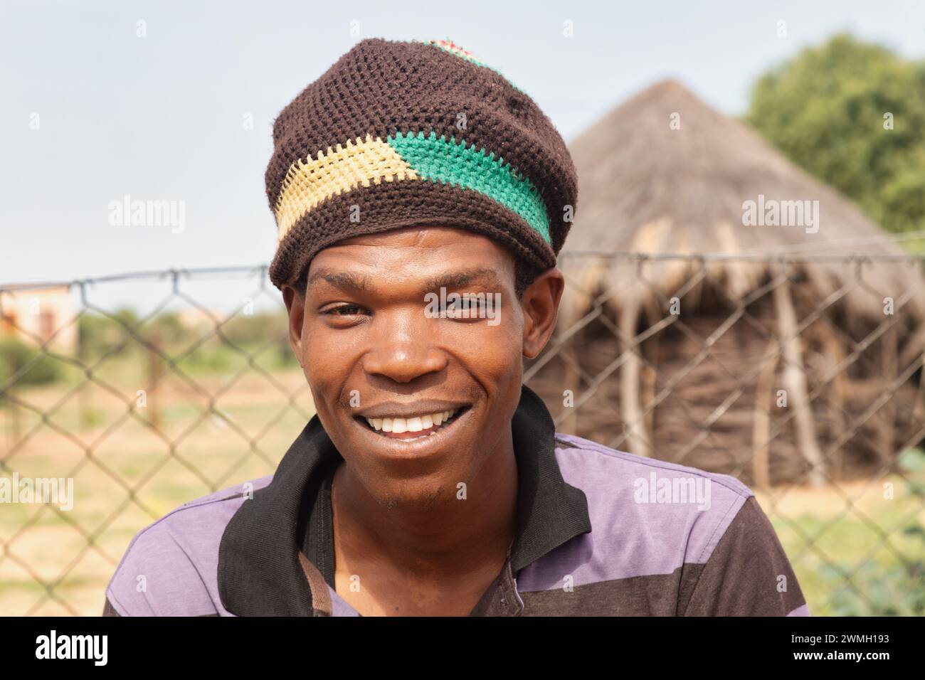 Junger afrikanischer Dorfmann mit einer Mütze, die vor einer strohgedeckten Hütte steht. Stockfoto