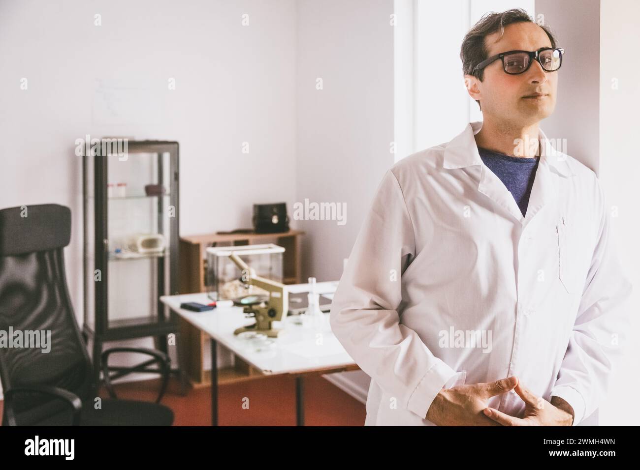 Ein erwachsener kaukasischer Wissenschaftler in Brille und weißem Mantel steht und blickt unermüdlich vor dem Hintergrund des Labors nach vorne. Stockfoto
