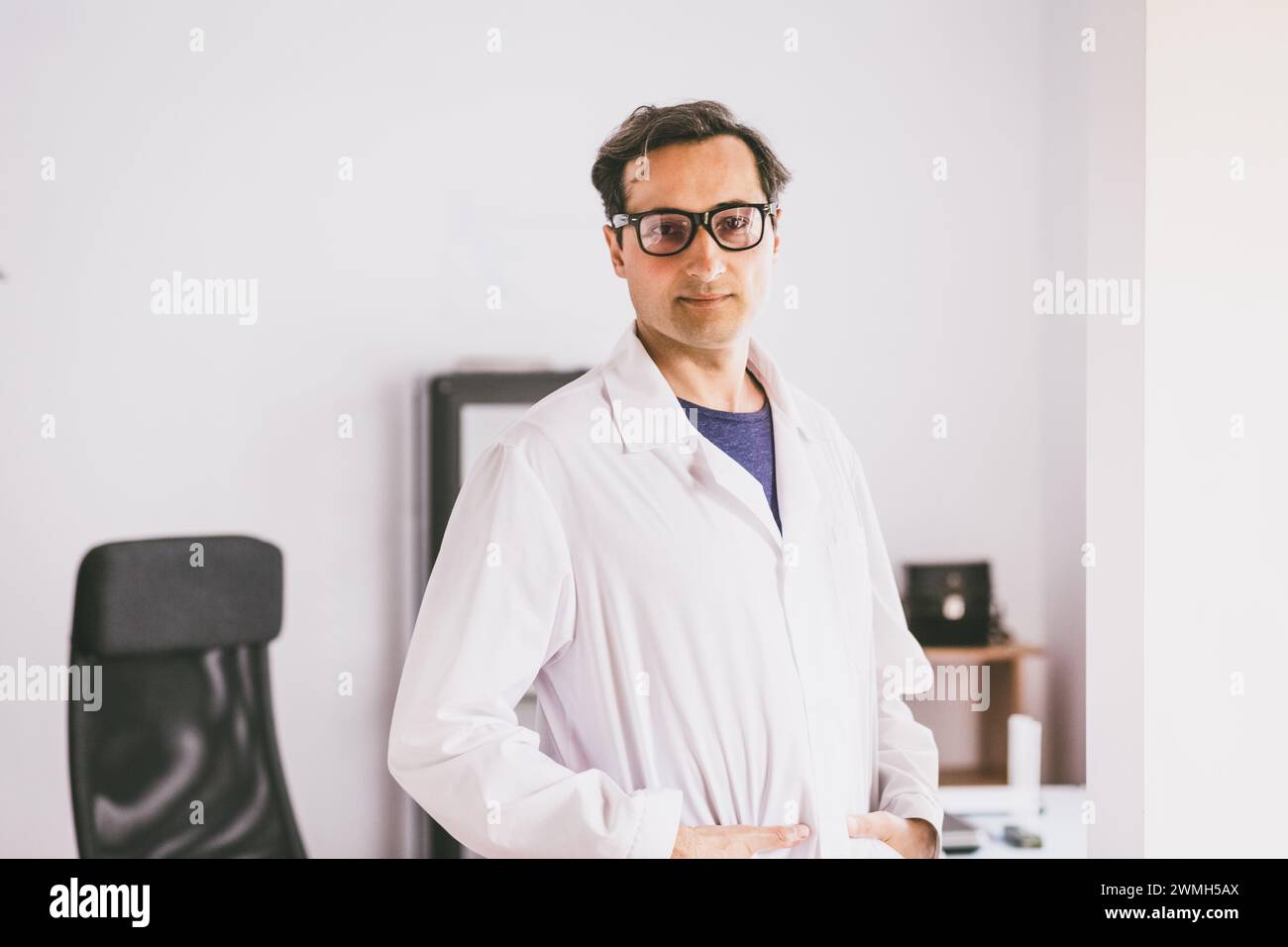 Ein erwachsener kaukasischer Wissenschaftler in Brille und weißem Mantel steht und blickt selbstbewusst vor dem Hintergrund des Labors nach vorne. Stockfoto