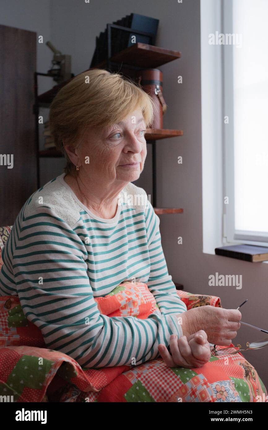 Eine ältere kaukasische Frau sitzt in einem Stuhl unter einer Decke am Fenster mit Brille in den Händen und ist in Gedanken verloren. Vertikale Zusammensetzung. Stockfoto