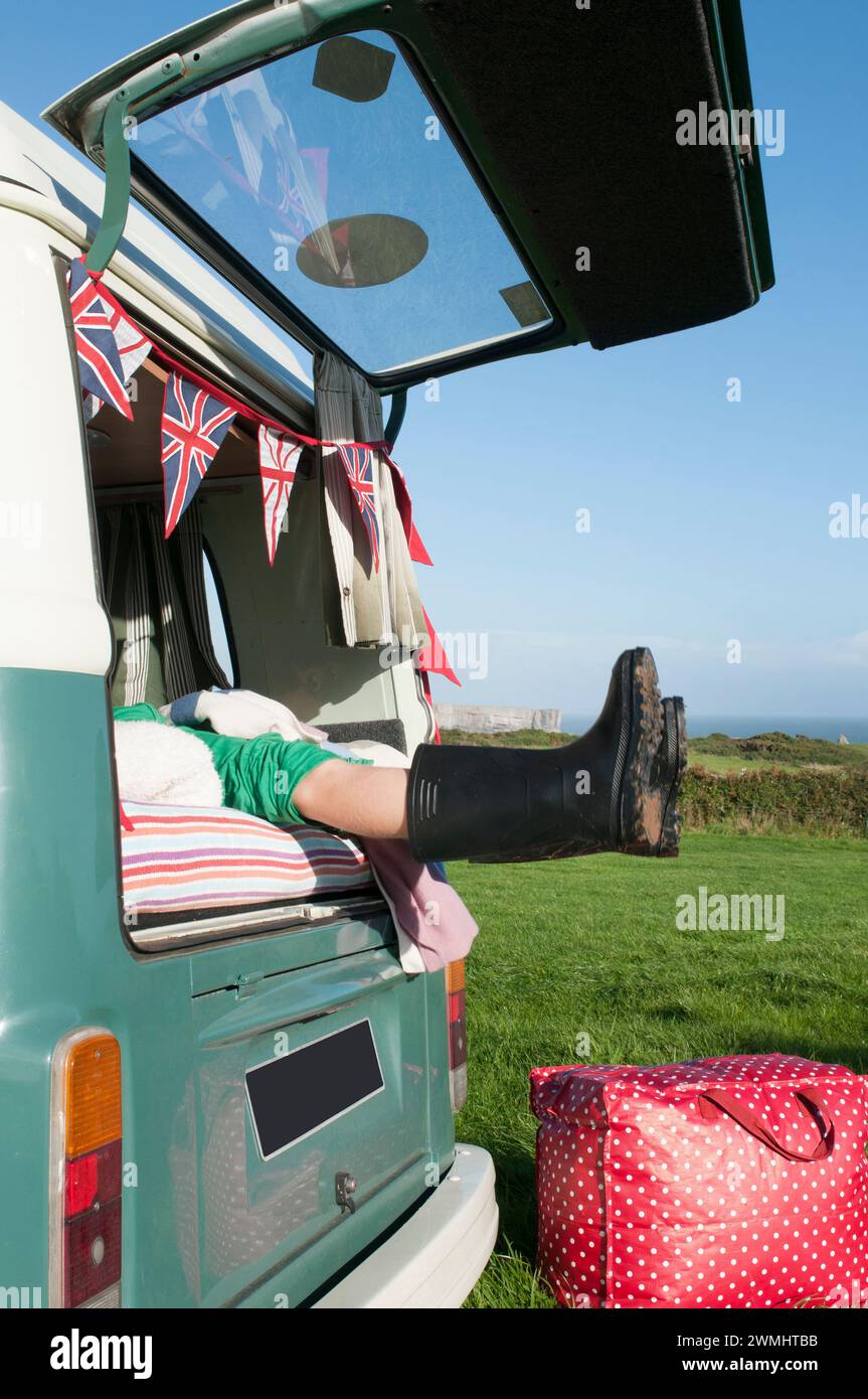 Ein Junge in Gummistiefeln auf der Rückseite eines alten Wohnwagens an einem sonnigen Tag. Stockfoto