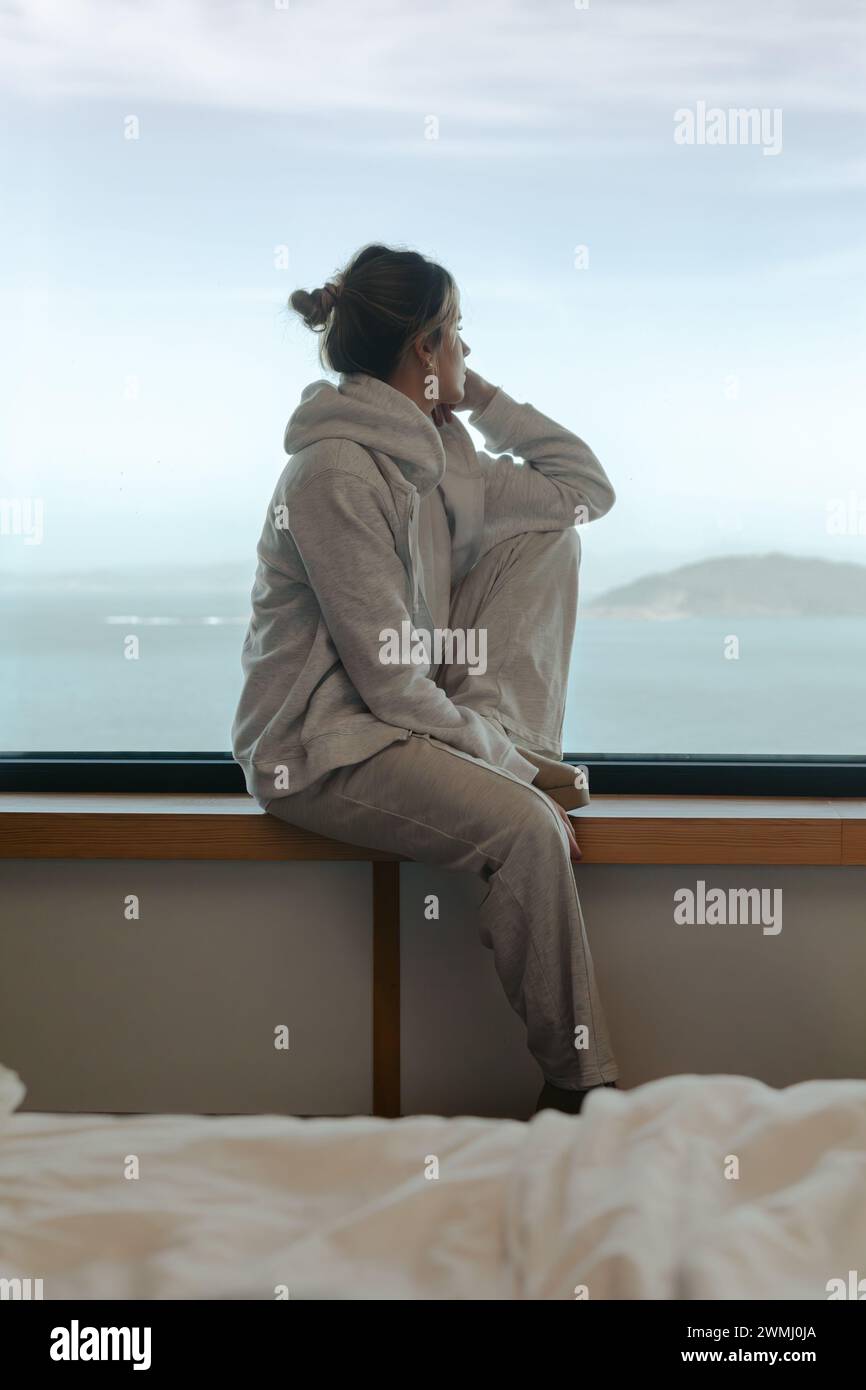 Besinnliche junge Frau, die vom Fenster eines modernen Zimmers aus in bequemer Loungewear auf das Meer blickt Stockfoto