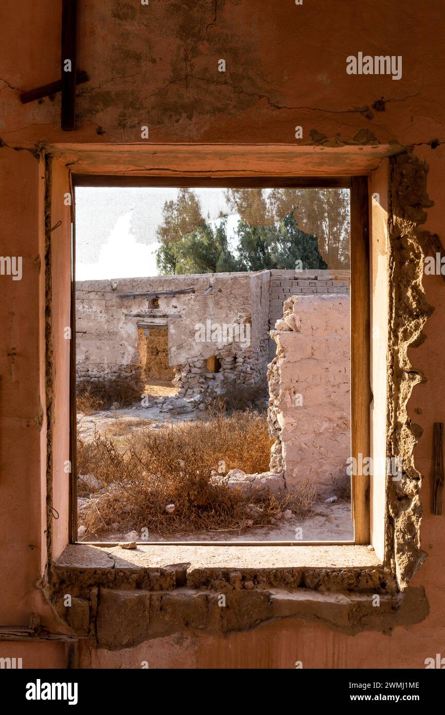 Blick durch ein zerbrochenes Fenster auf ruinierte Gebäude und Innenhof in Al Dschazirah Al Hamra Spukstadt in Ras Al Khaimah, Vereinigte Arabische Emirate. Stockfoto