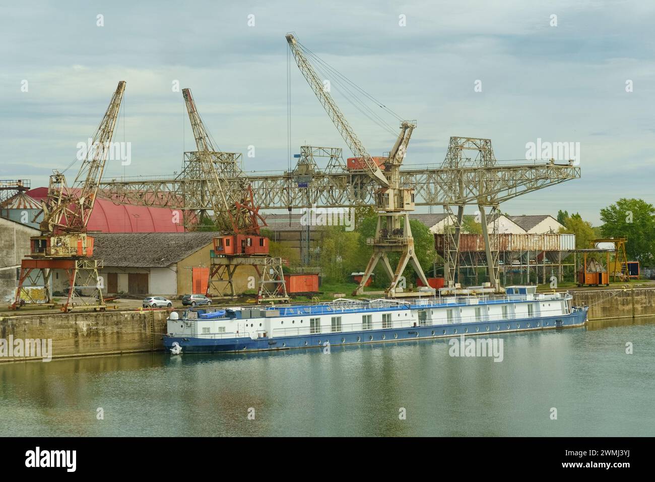 Saint-Marseille, Frankreich, 28. April 2023: Ein blauer Frachtkahn schwimmt auf einem ruhigen Fluss, der an einem Industriehafen angedockt ist. Dahinter, große Metallkräne und Wa Stockfoto