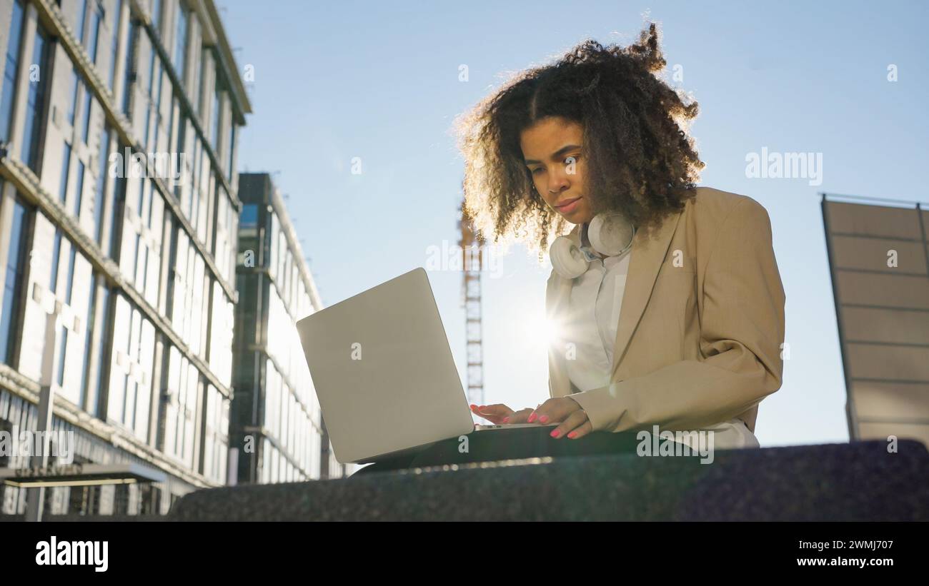 Junge professionelle Frau konzentriert sich auf ihren Laptop-Bildschirm, während sie draußen arbeitet, mit modernen Gebäuden im Hintergrund. Stockfoto