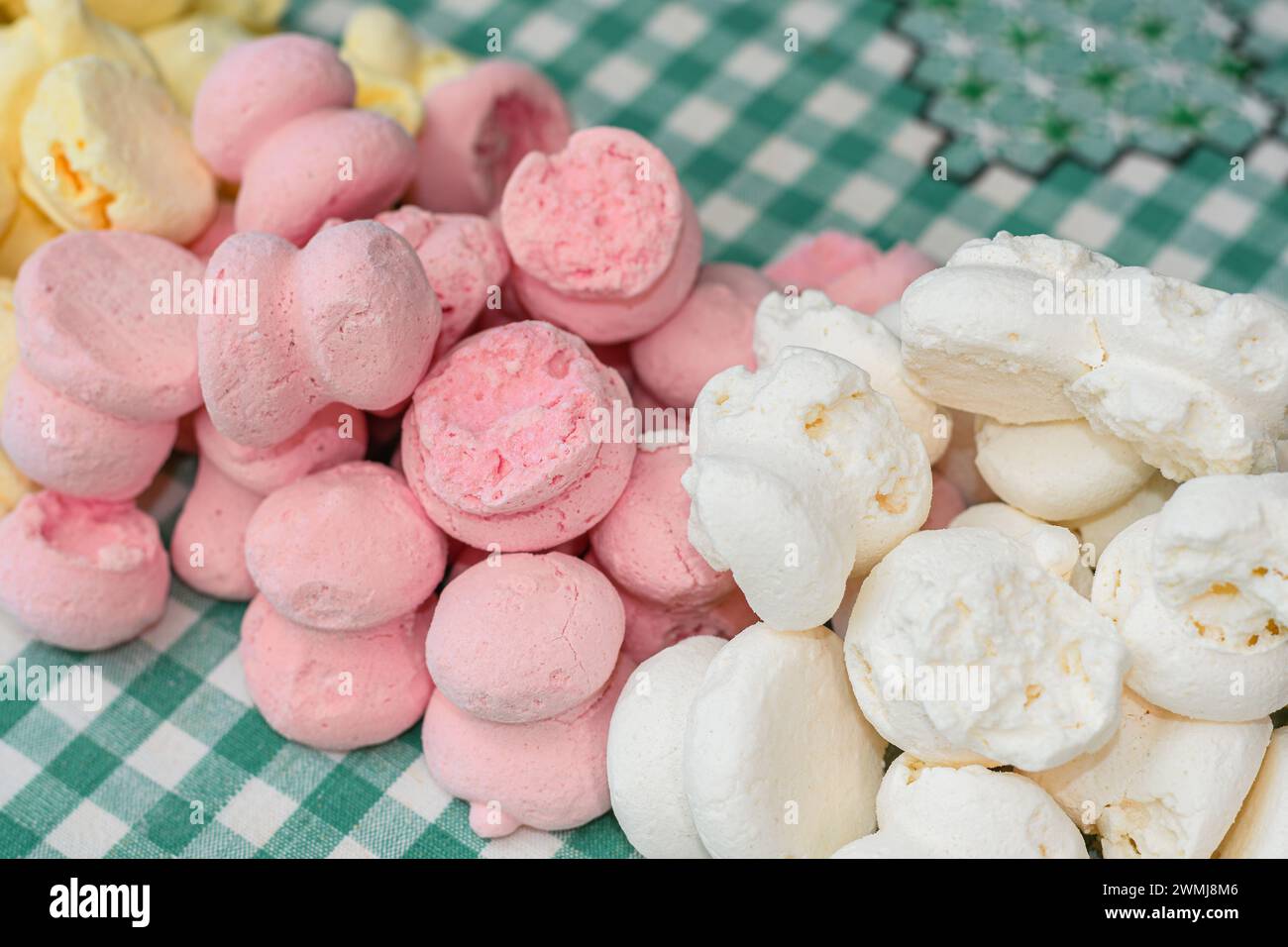 Hintergrund von farbigen hausgemachten Meringue-Bonbons über einer Tischdecke mit rustikaler karierter Textur. Gelbe, rosa und weiße Meringue, brasilianische Süßigkeiten. Stockfoto