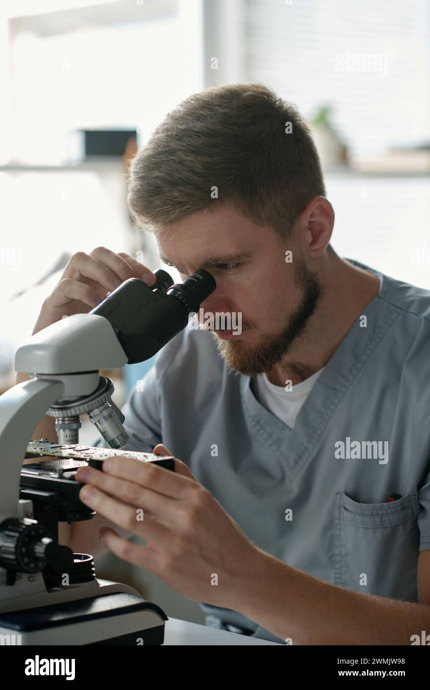 Junger seriöser männlicher Techniker des Fehlerbehebungszentrums, der auf das Motherboard im Mikroskop blickt, während er am Arbeitsplatz sitzt Stockfoto