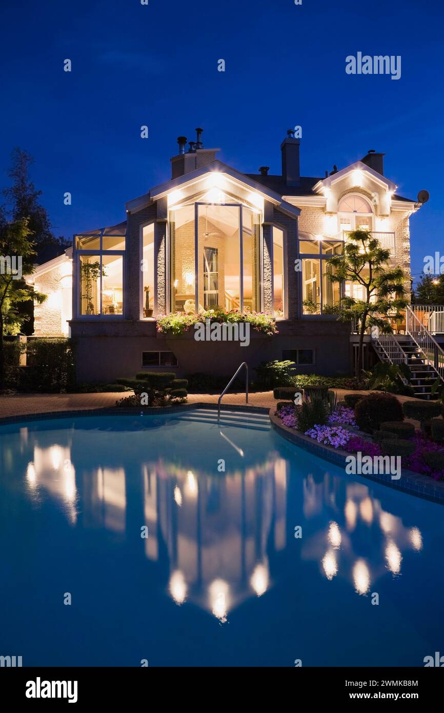 Modernes Haus mit großen Panoramafenstern aus Glas und landschaftlich gestaltetem Hinterhof mit Pool in der Dämmerung. Stockfoto