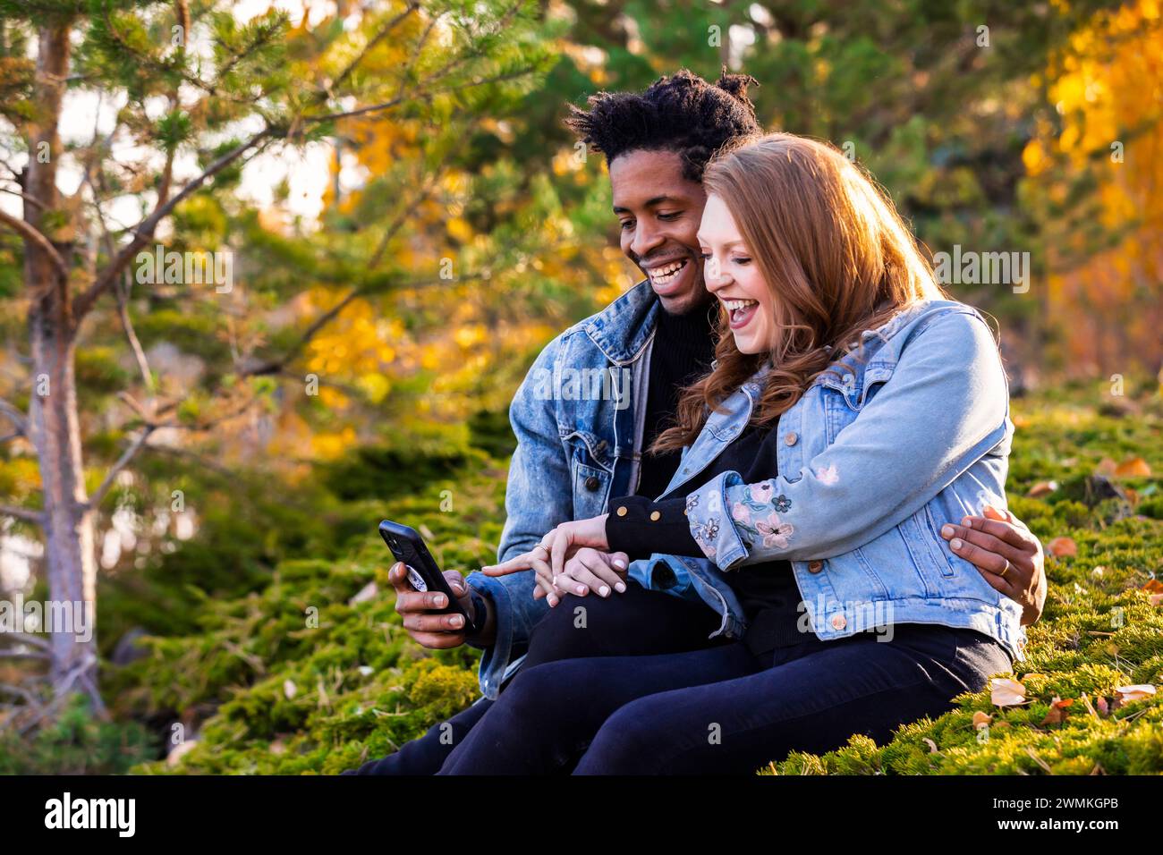 Nahaufnahme einer gemischten Rasse verheiratetes Paar, das lächelt und zusammen auf das Handy schaut, in der Dämmerung auf dem Gras sitzt, schöne Zeit miteinander verbringt... Stockfoto