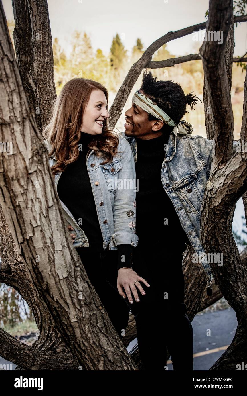 Nahaufnahme eines Paares gemischter Rassen, das sich anlächelt, während er sich auf Ästen ausruht und während einer Herbstfamilie ... gute Zeit miteinander verbringt Stockfoto