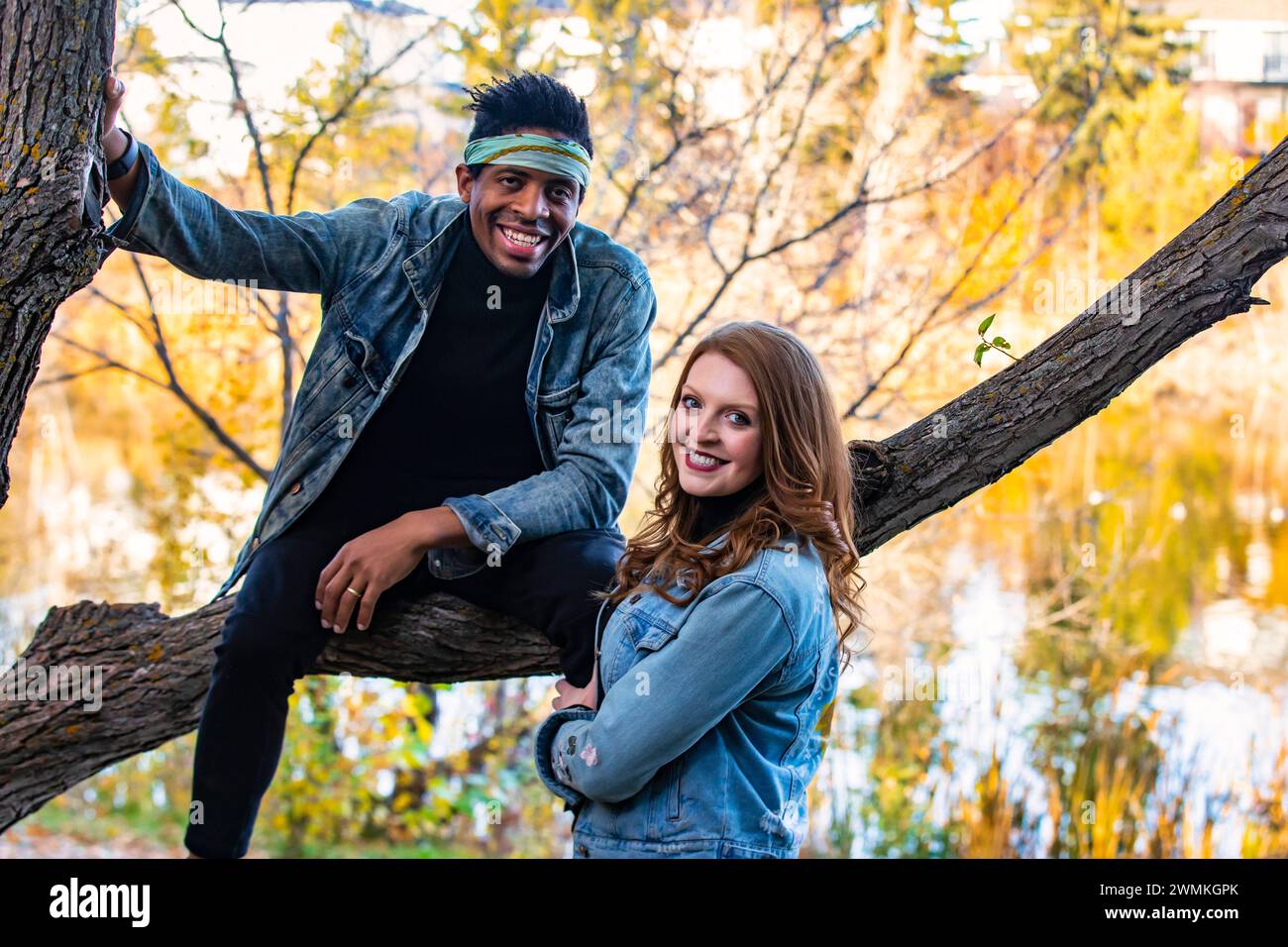 Nahaufnahme eines Paares gemischter Rassen, das auf einem Baumzweig in die Kamera lächelt und während einer Herbstfamilie ... wertvolle Zeit miteinander verbringt Stockfoto