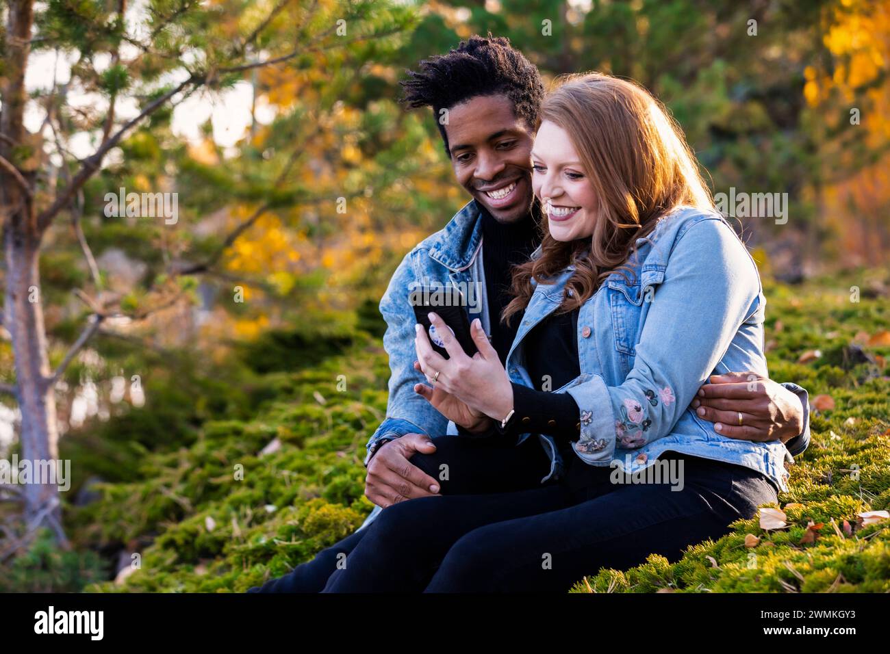 Nahaufnahme einer gemischten Rasse verheiratetes Paar, das lächelt und zusammen auf das Handy schaut, in der Dämmerung auf dem Gras sitzt, schöne Zeit miteinander verbringt... Stockfoto