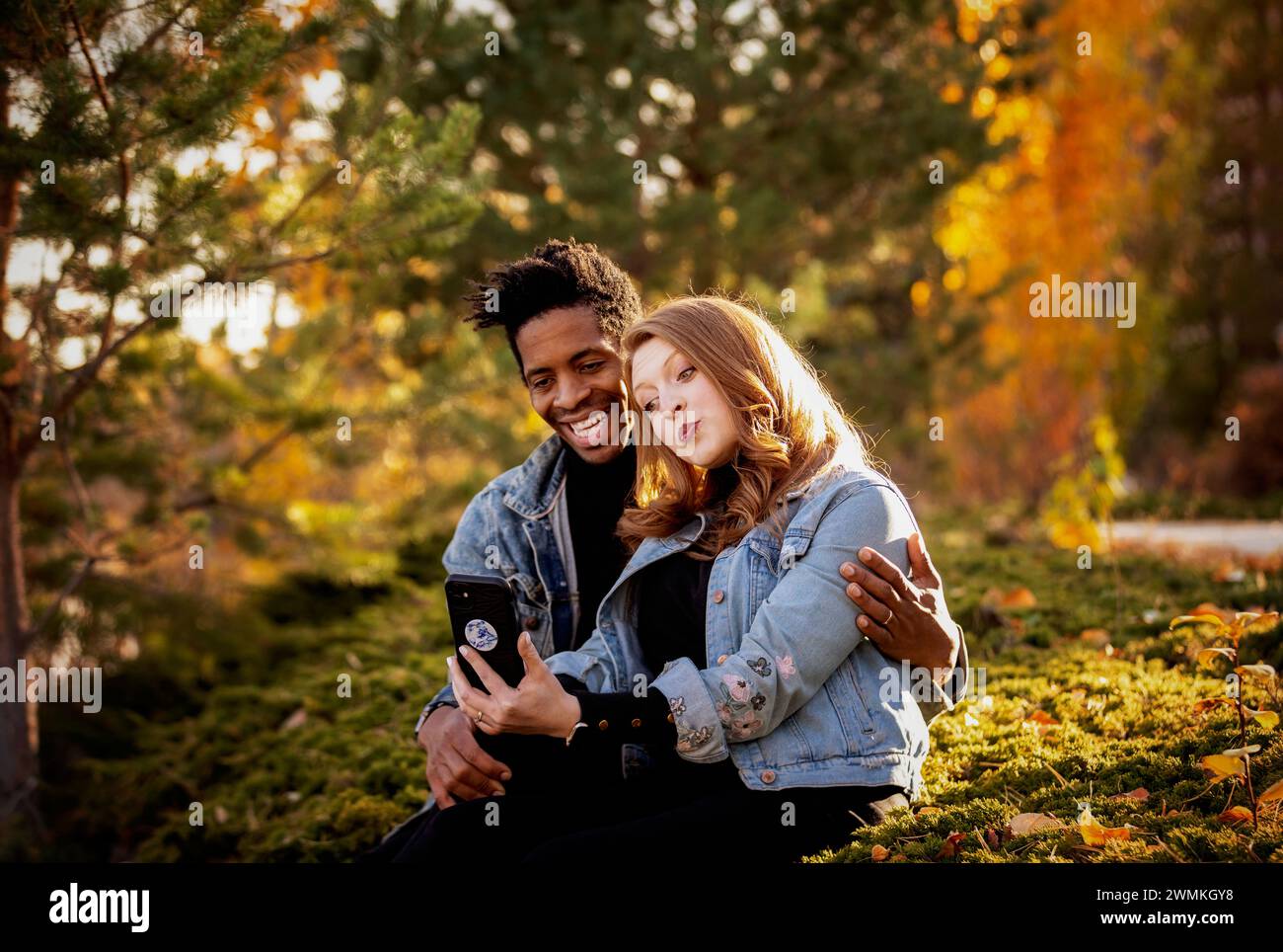 Nahaufnahme einer gemischten Rasse verheiratete Paare, die sich umarmen und Gesichter machen, während sie ein Selfie machen und in der Abenddämmerung auf dem Gras sitzen, Qualität verbringen tim... Stockfoto