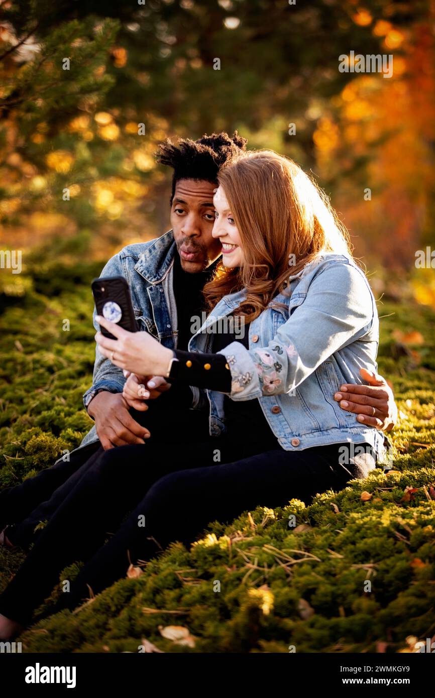 Nahaufnahme einer gemischten Rasse verheiratete Paare, die sich umarmen und Gesichter machen, während sie ein Selfie machen und in der Abenddämmerung auf dem Gras sitzen, Qualität verbringen tim... Stockfoto
