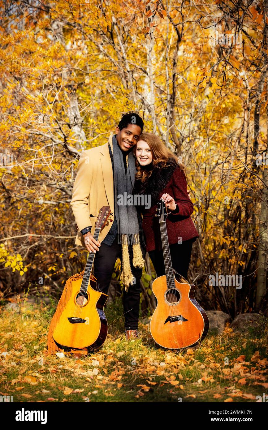 Porträt eines gemischten Ehepaares mit Akustikgitarren und einem Lächeln in die Kamera, während sie während einer Herbstfamilie Zeit miteinander verbringen... Stockfoto