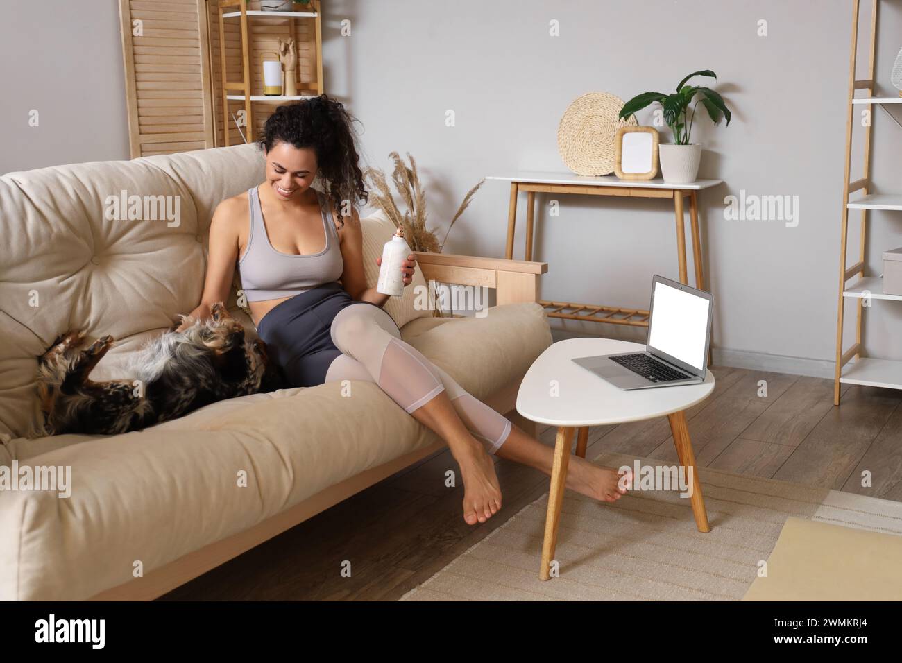 Sportliche junge afroamerikanische Frau mit Wasserflasche und Cocker Spaniel, die zu Hause auf dem Sofa sitzt Stockfoto