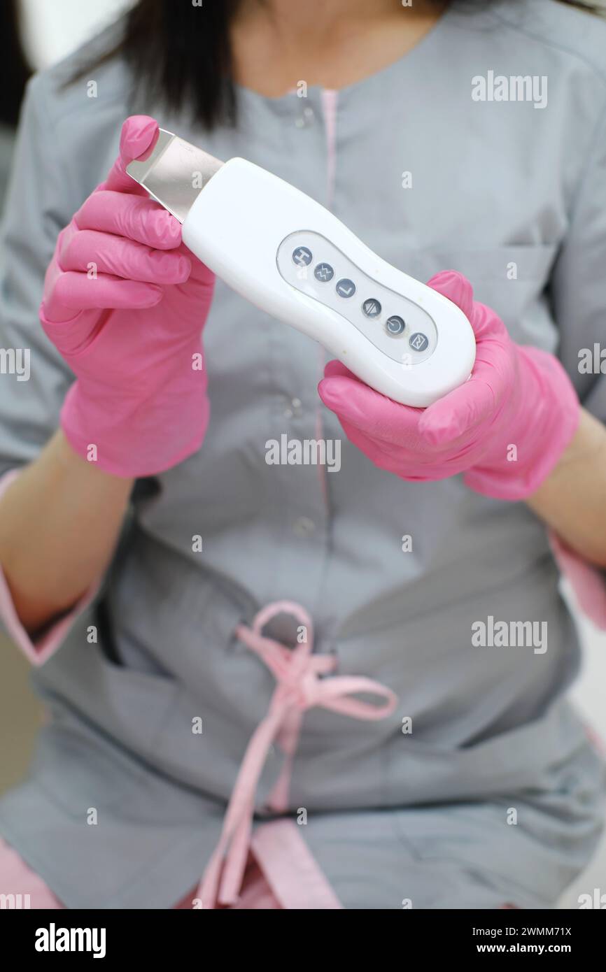 Ein Kosmetiker in pinkfarbenen Handschuhen zeigt ein weißes Ultraschall-Hautpflegegerät mit Funktionstasten. Vertikales Foto Stockfoto